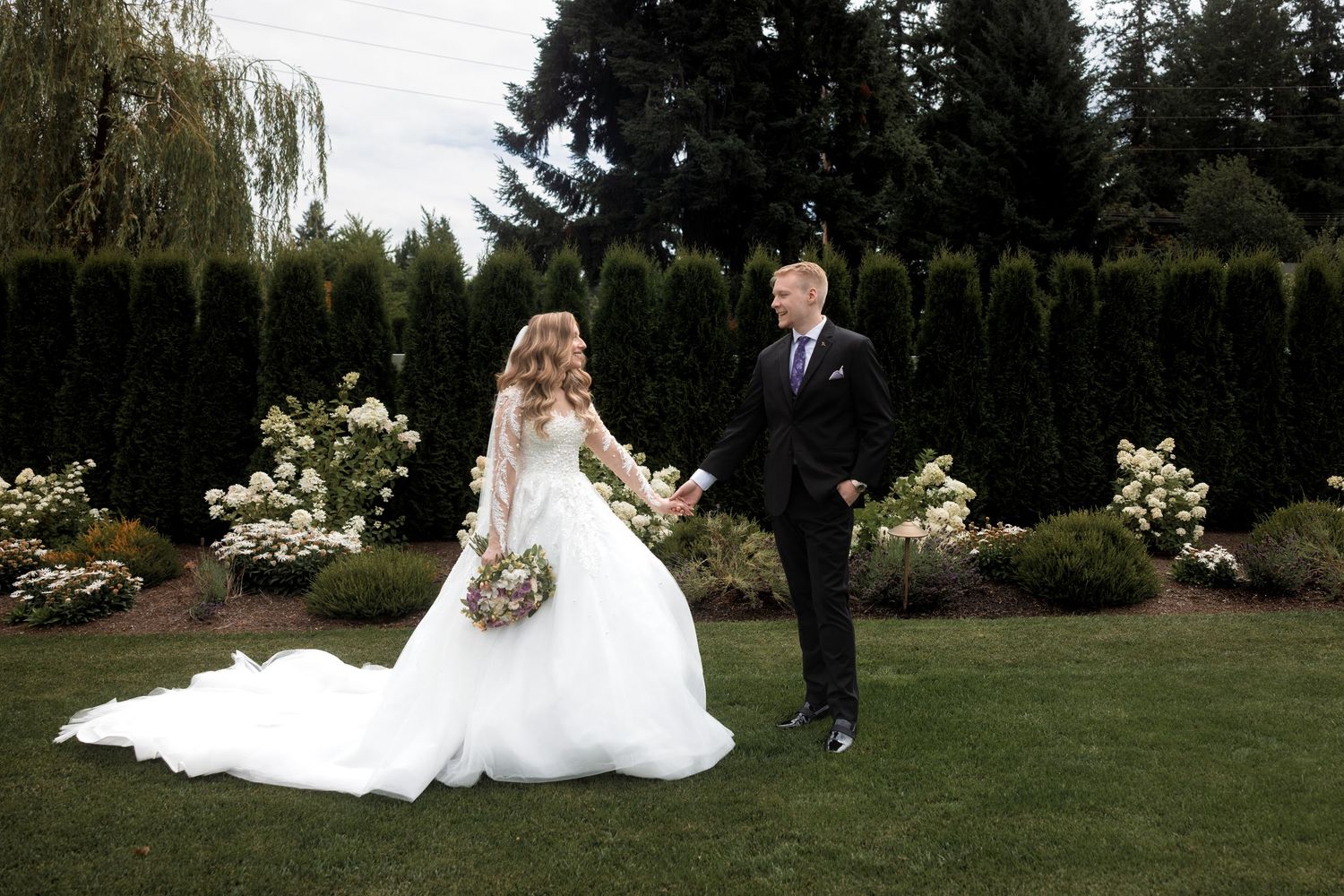 Wedding couple walking through manicured garden with evergreen backdrop.
