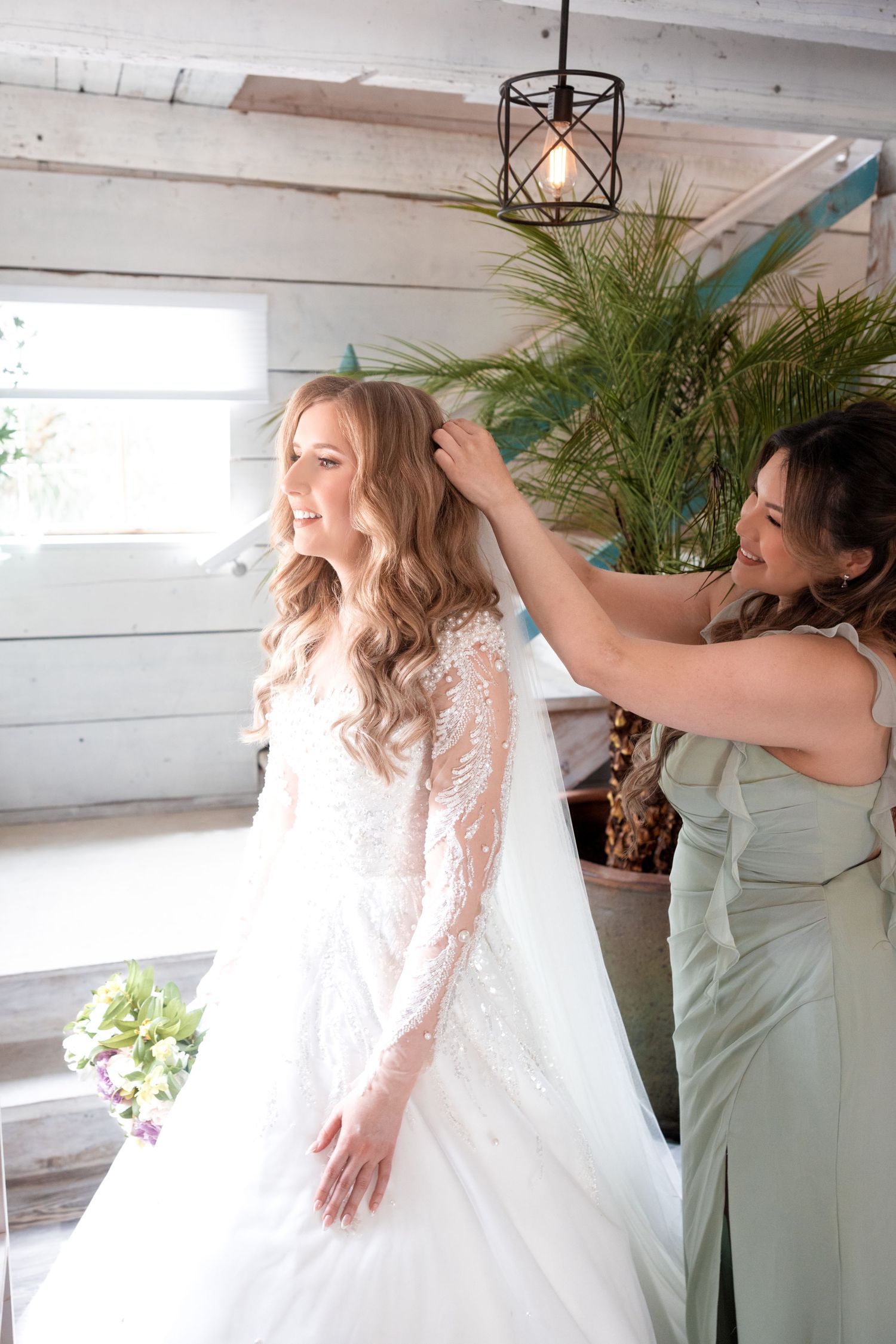 Getting ready scene shows final dress adjustments in a bright indoor space with tropical plants.