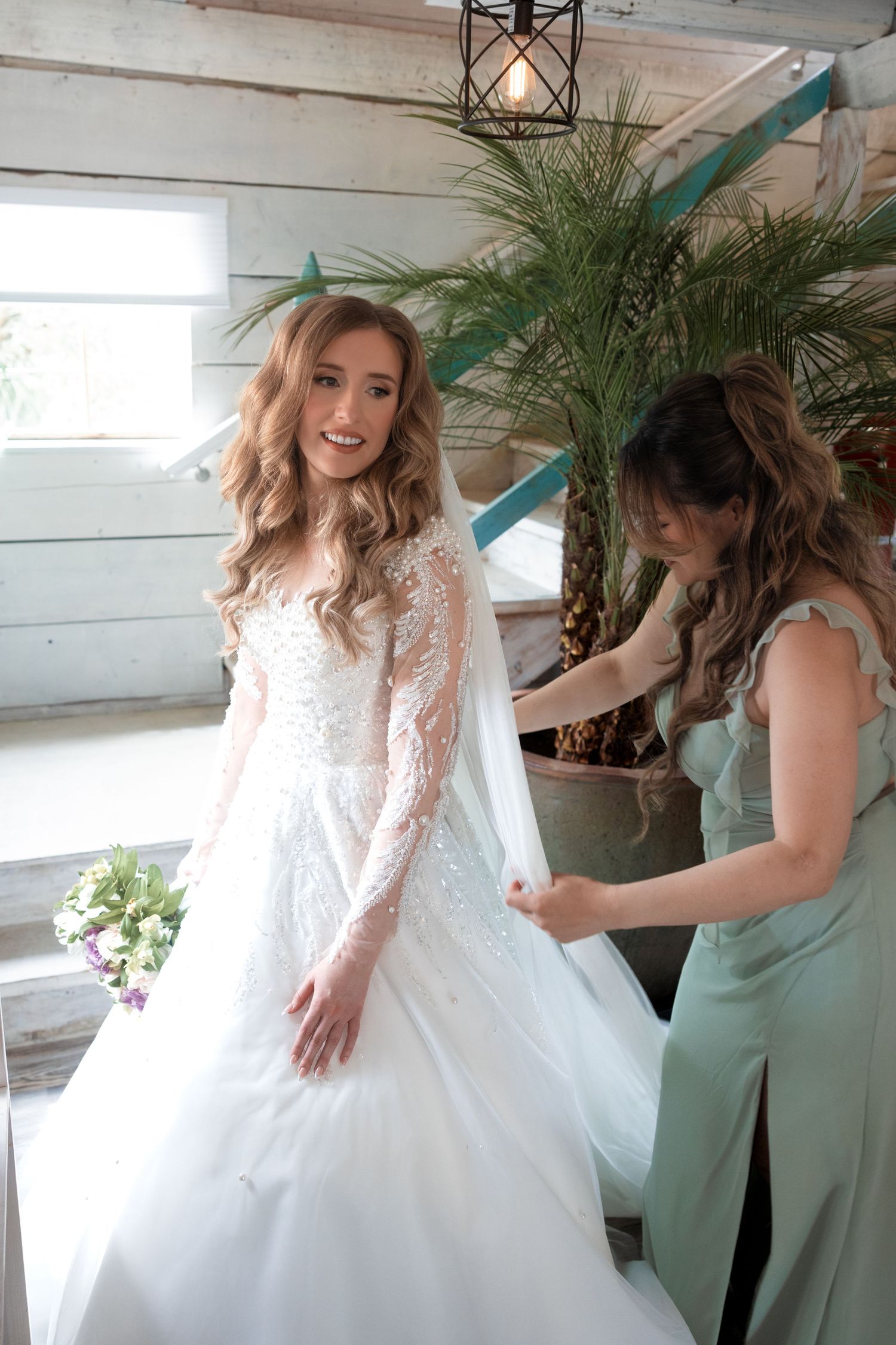 A bride gets help with her wedding dress fitting in a rustic room with greenery.