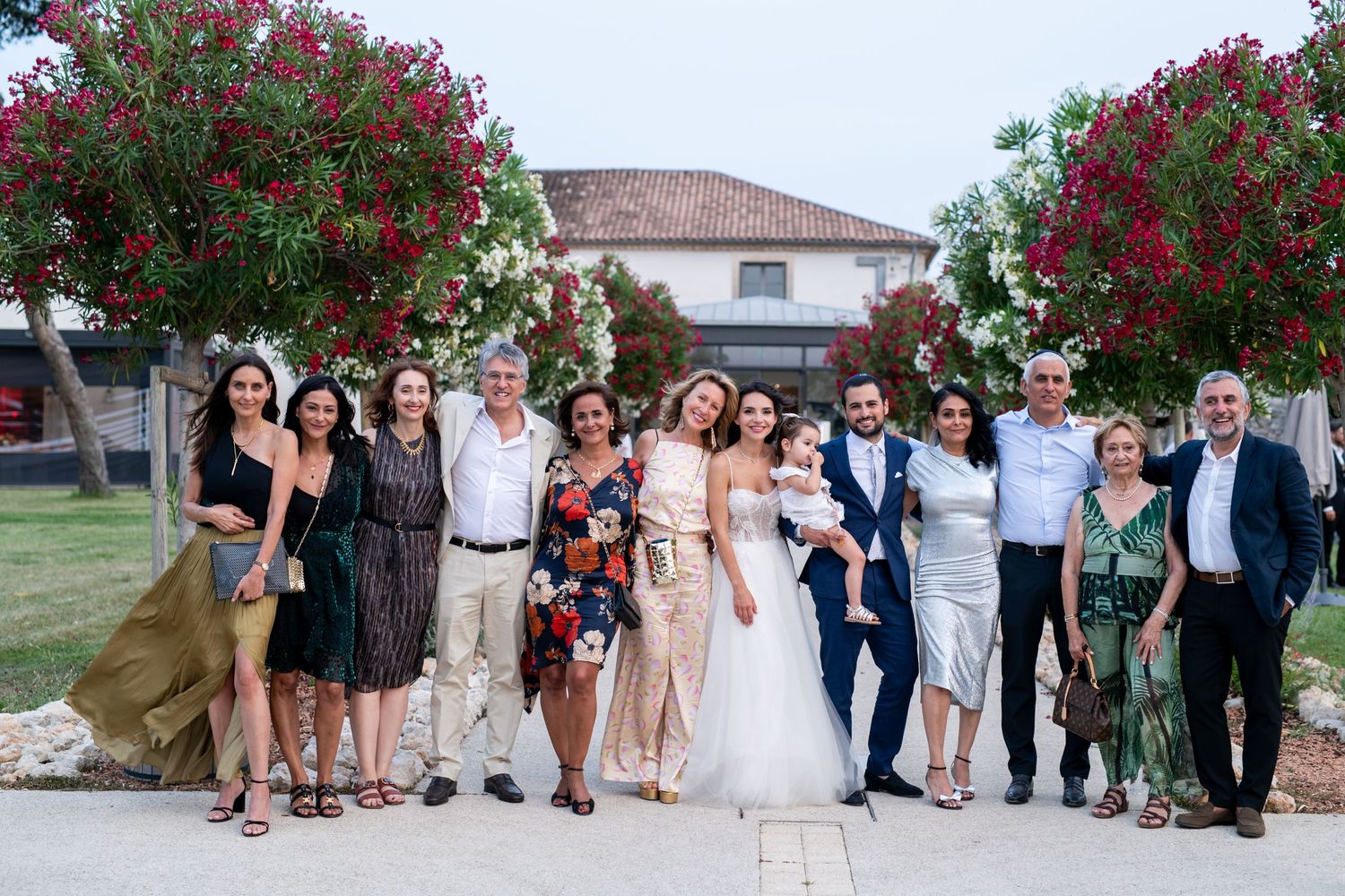 Photo de groupe du mariage devant un bâtiment fleuri, famille et invités réunis dans une ambiance estivale.