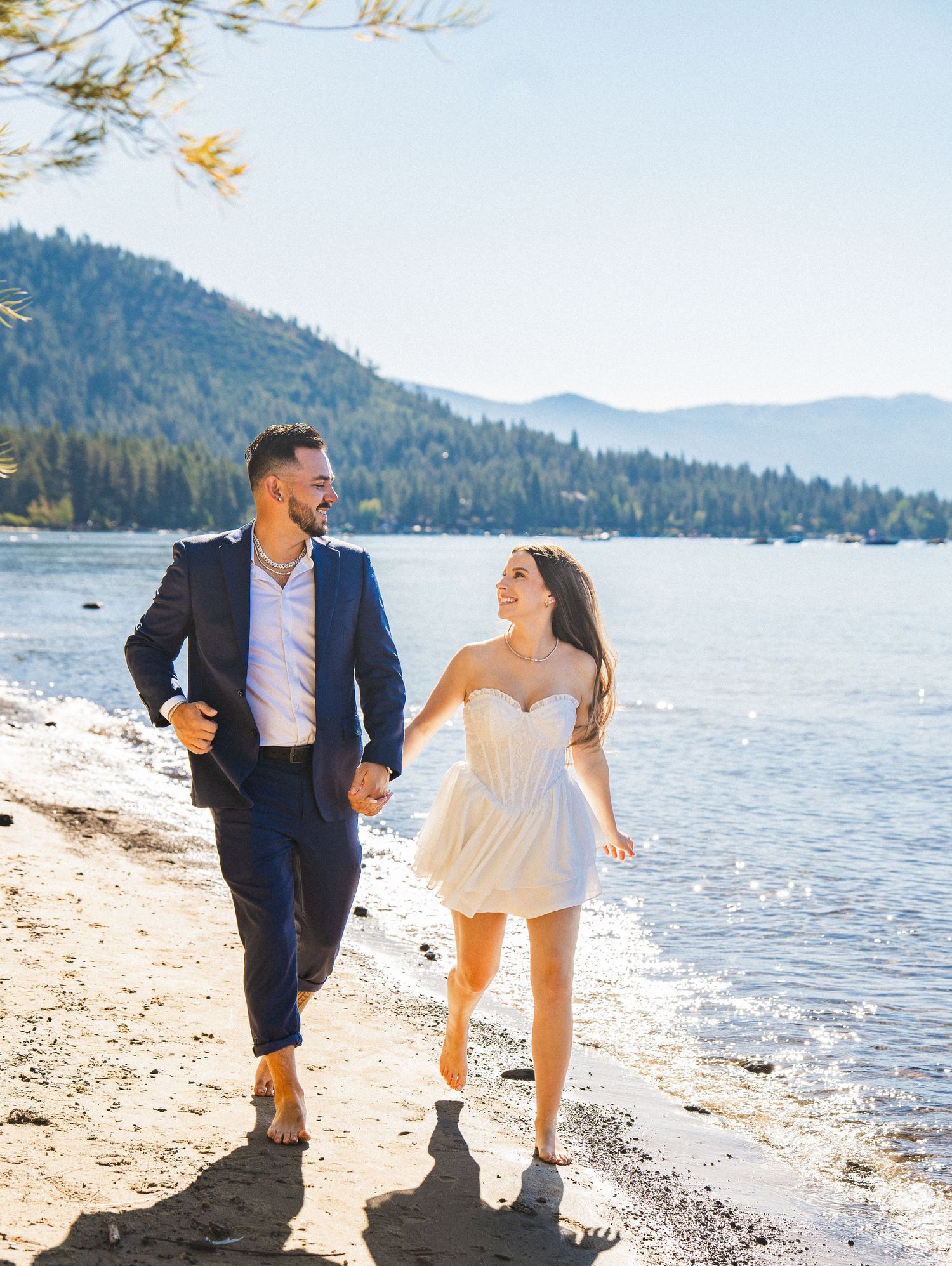 Couple enjoying a romantic walk along a scenic lakeside beach with mountains in the background.