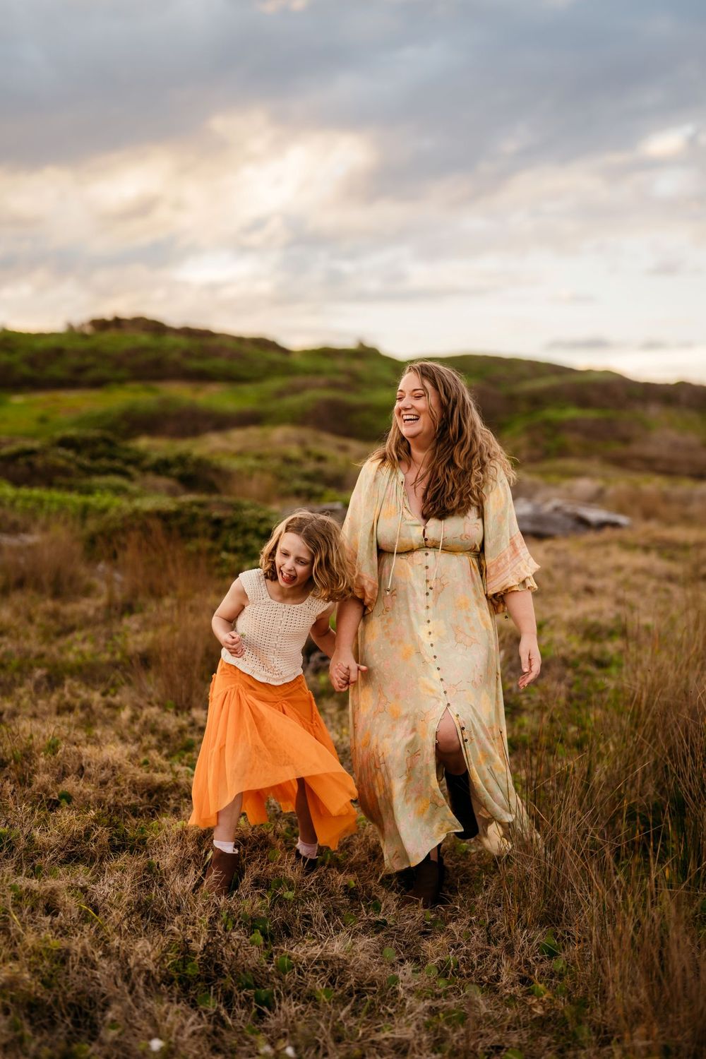 mother and child in flowing dresses walk together and laugh while exploring a grassy hillside at sunset.