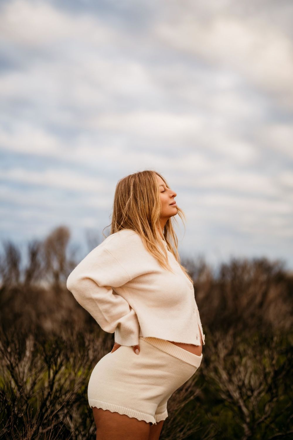 A pregnant woman in cream coloured clothing poses expressively against a dark treeline and cloudy sky background.