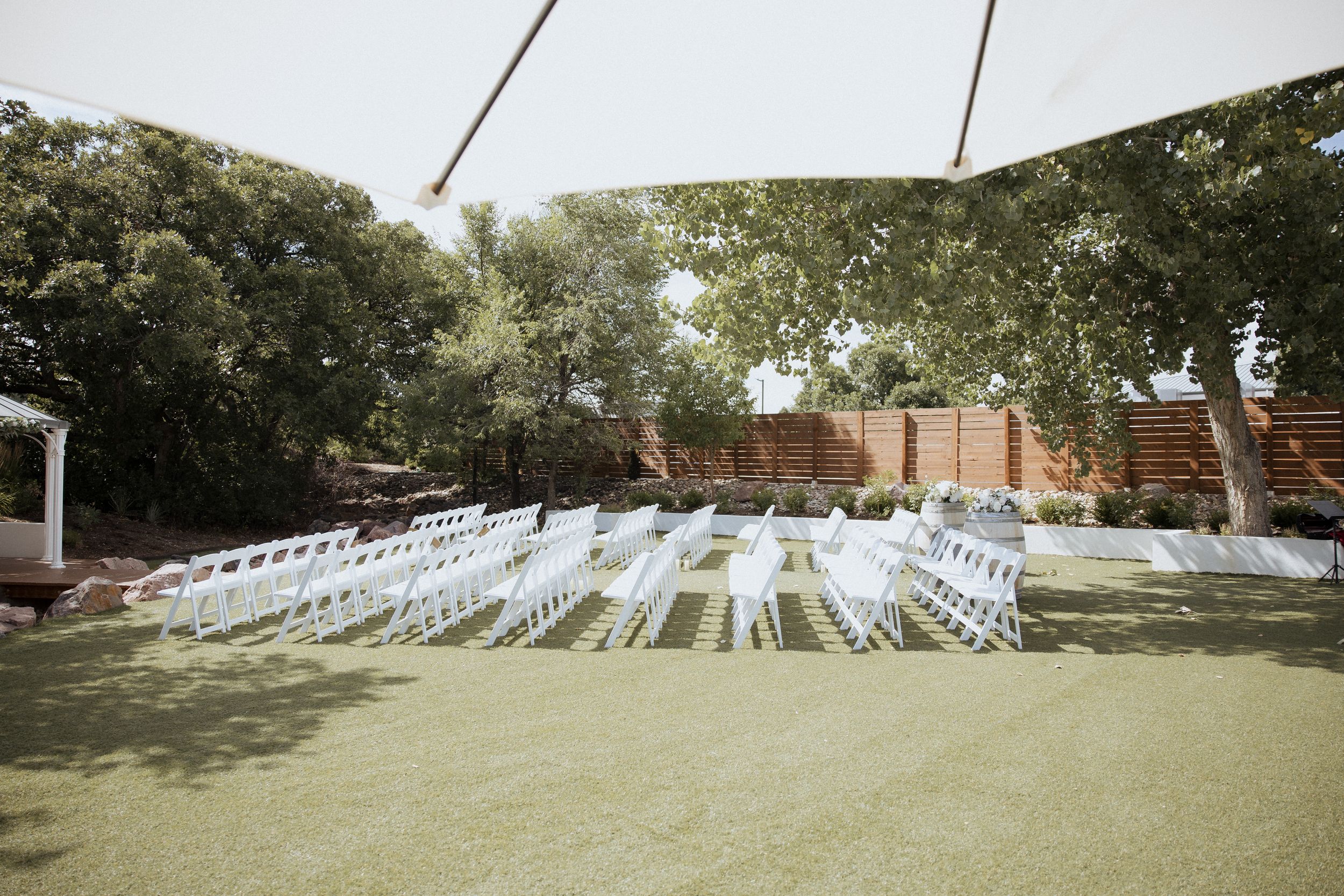 White wedding chairs arranged in rows on a grassy lawn under shady trees for an outdoor ceremony.