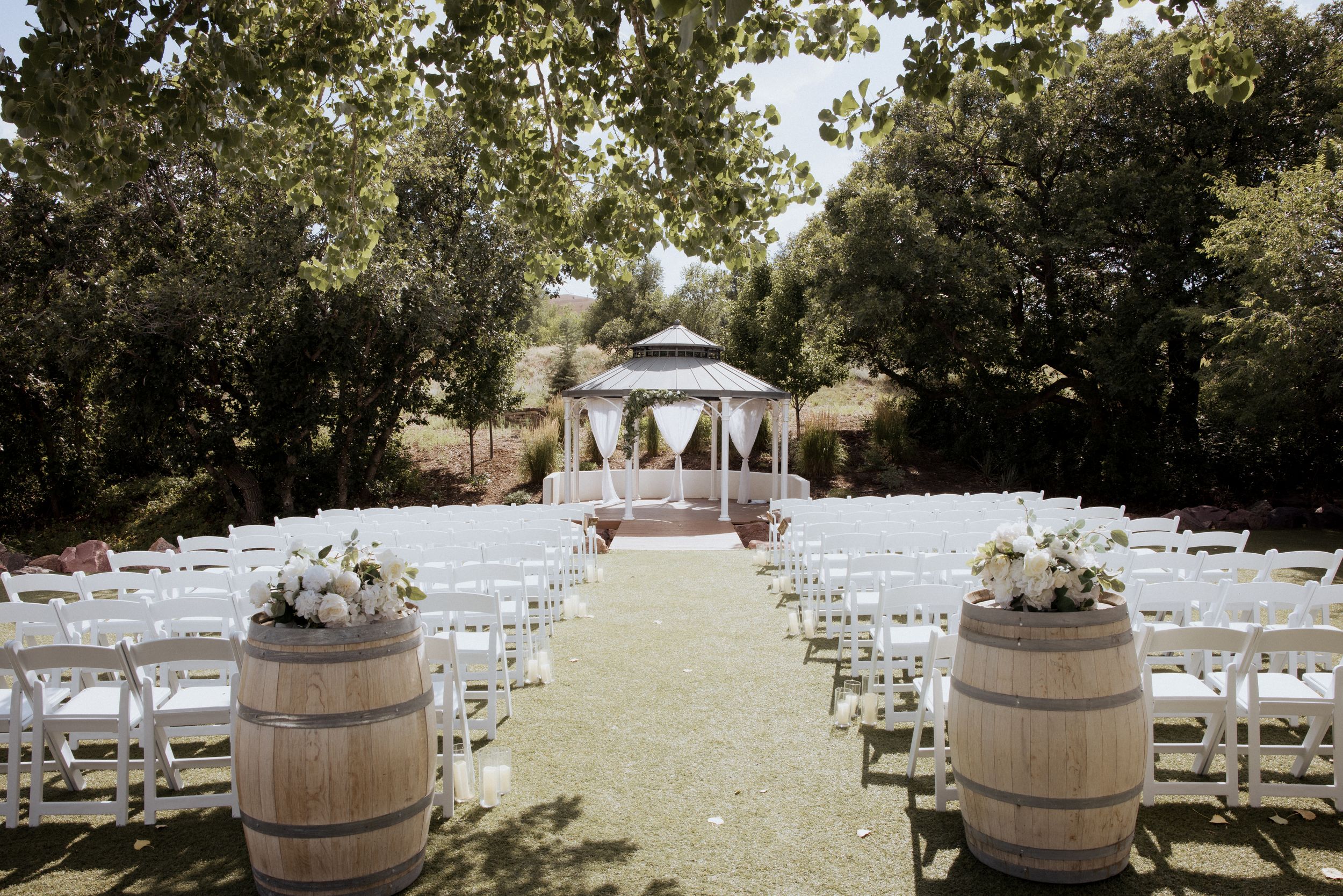 Rustic outdoor wedding ceremony setup with wooden barrels and white chairs arranged on a grassy lawn.