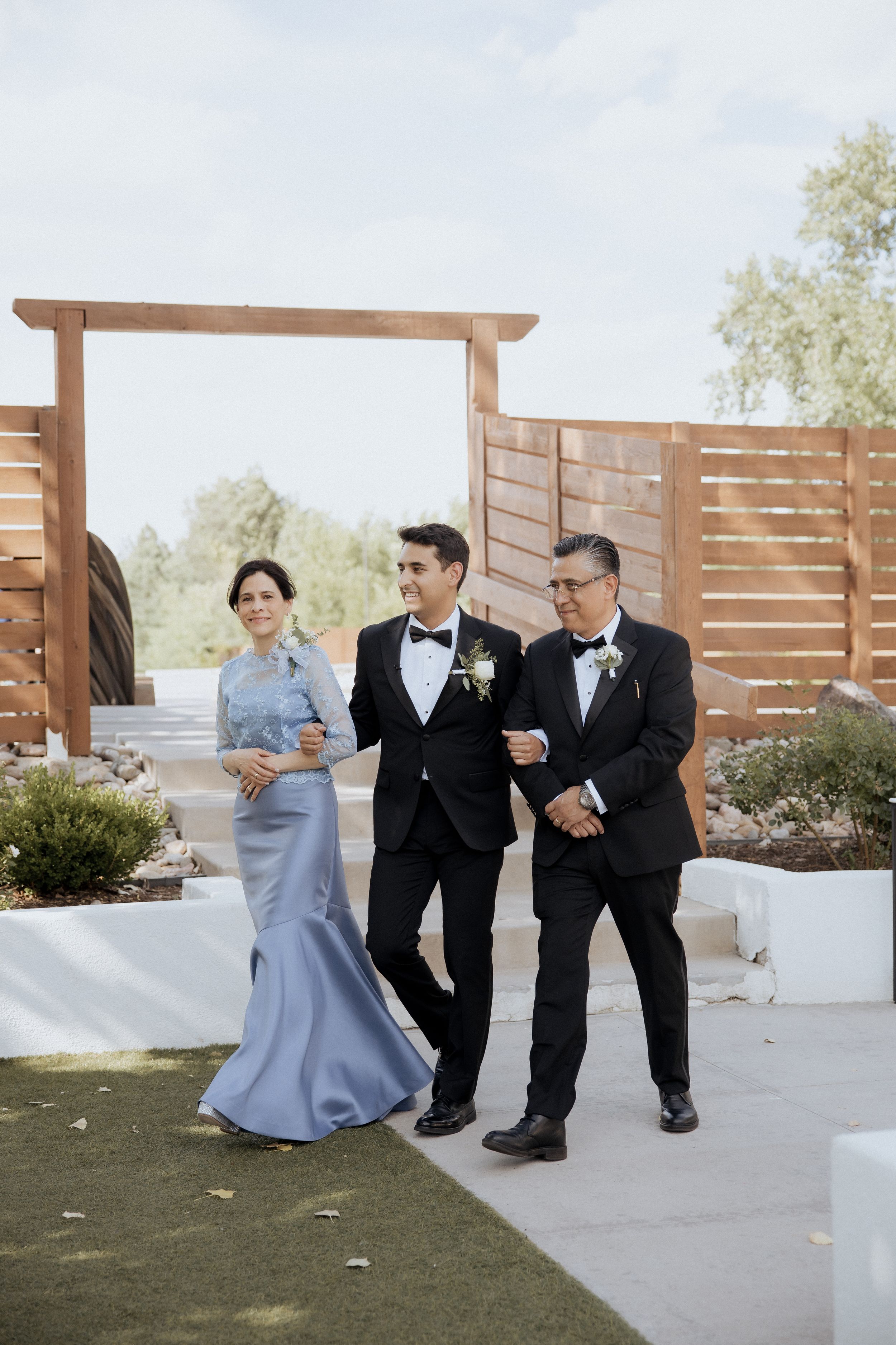 Three people in formal attire sit and laugh together on white steps with wooden privacy screens in the background.
