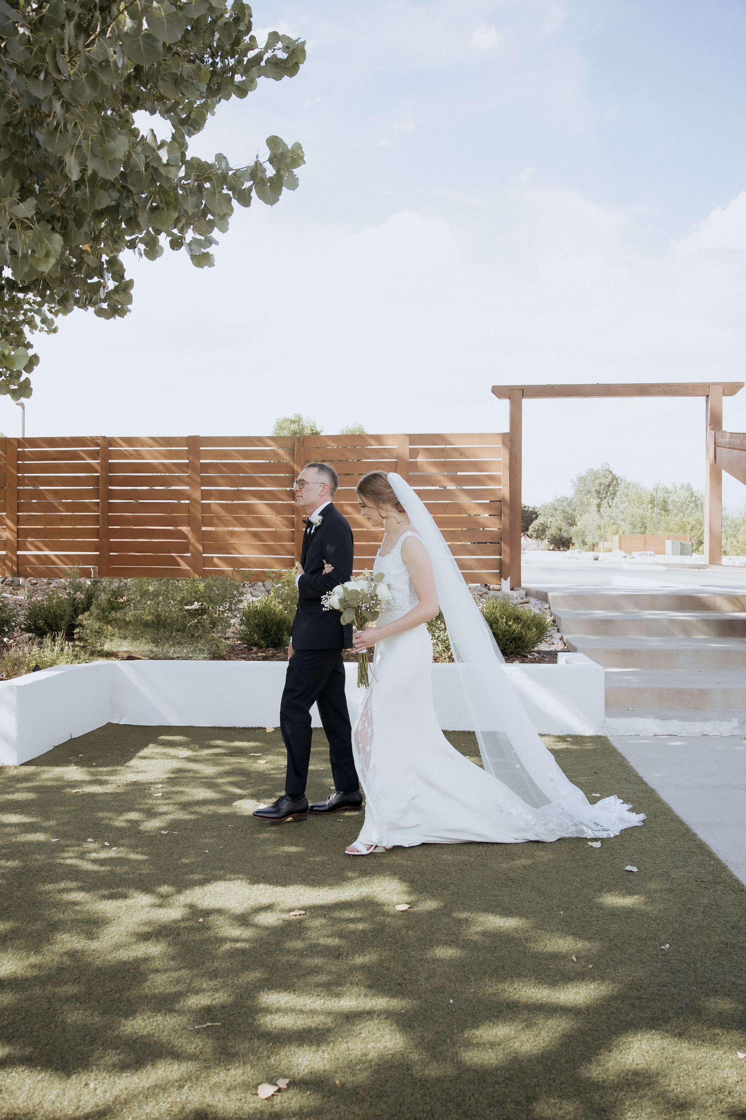 A romantic first look moment between a couple in formal wedding attire on a sunny outdoor terrace with wooden privacy walls.