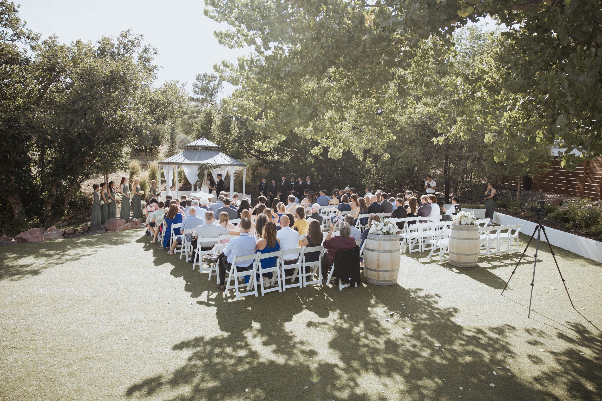 A row of white chairs set up for an outdoor wedding ceremony under shaded trees on a sunny day.