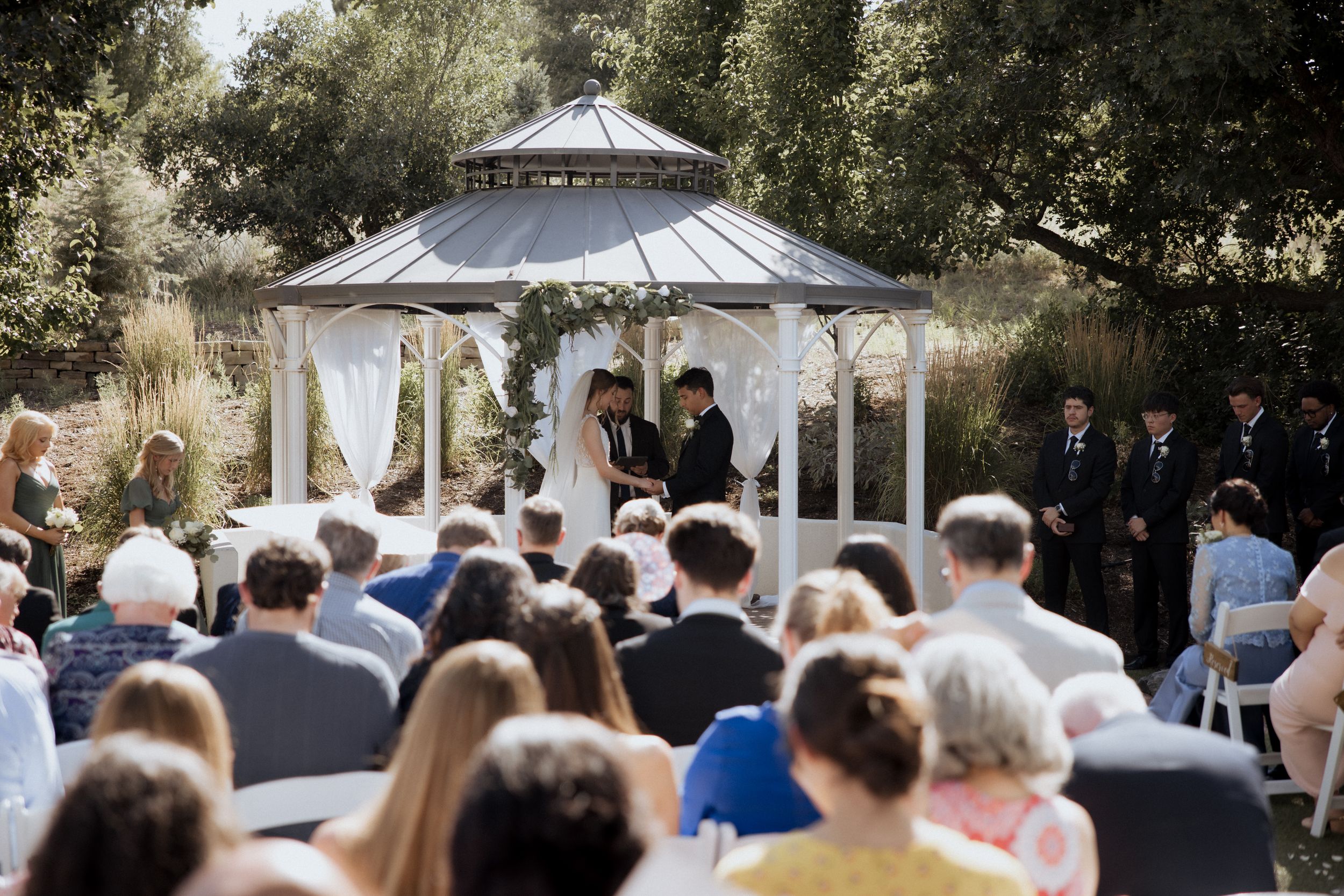 An outdoor wedding ceremony takes place under a white gazebo surrounded by trees and guests seated on white chairs.