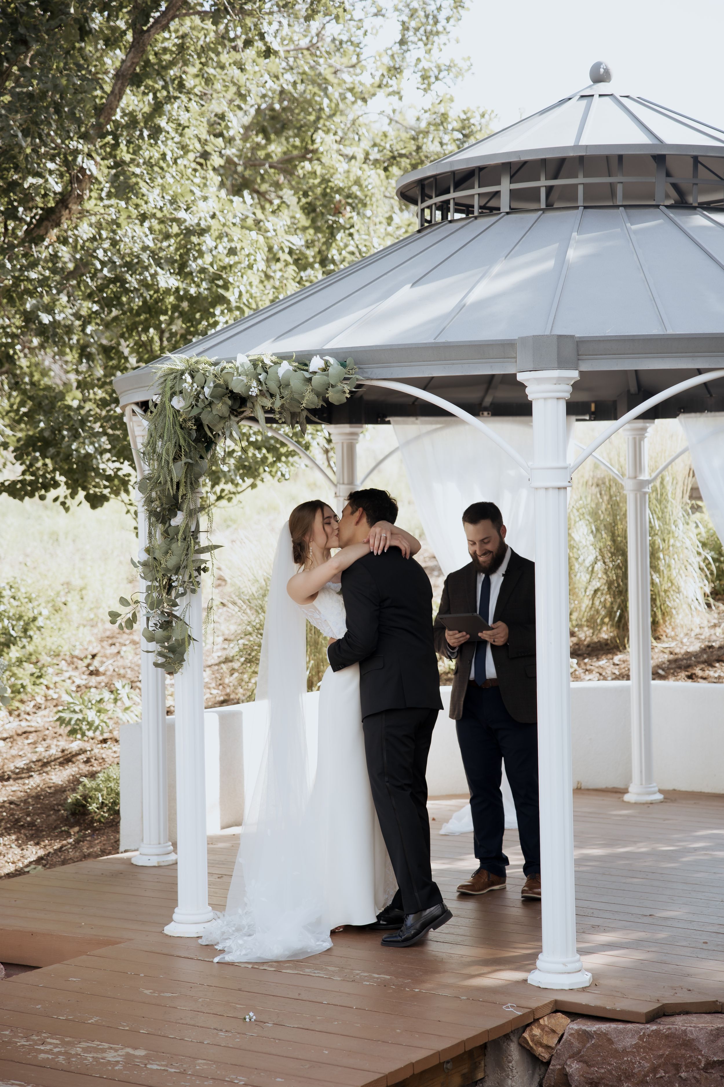 A romantic wedding ceremony unfolds under a white gazebo with elegant columns and a wooden deck.