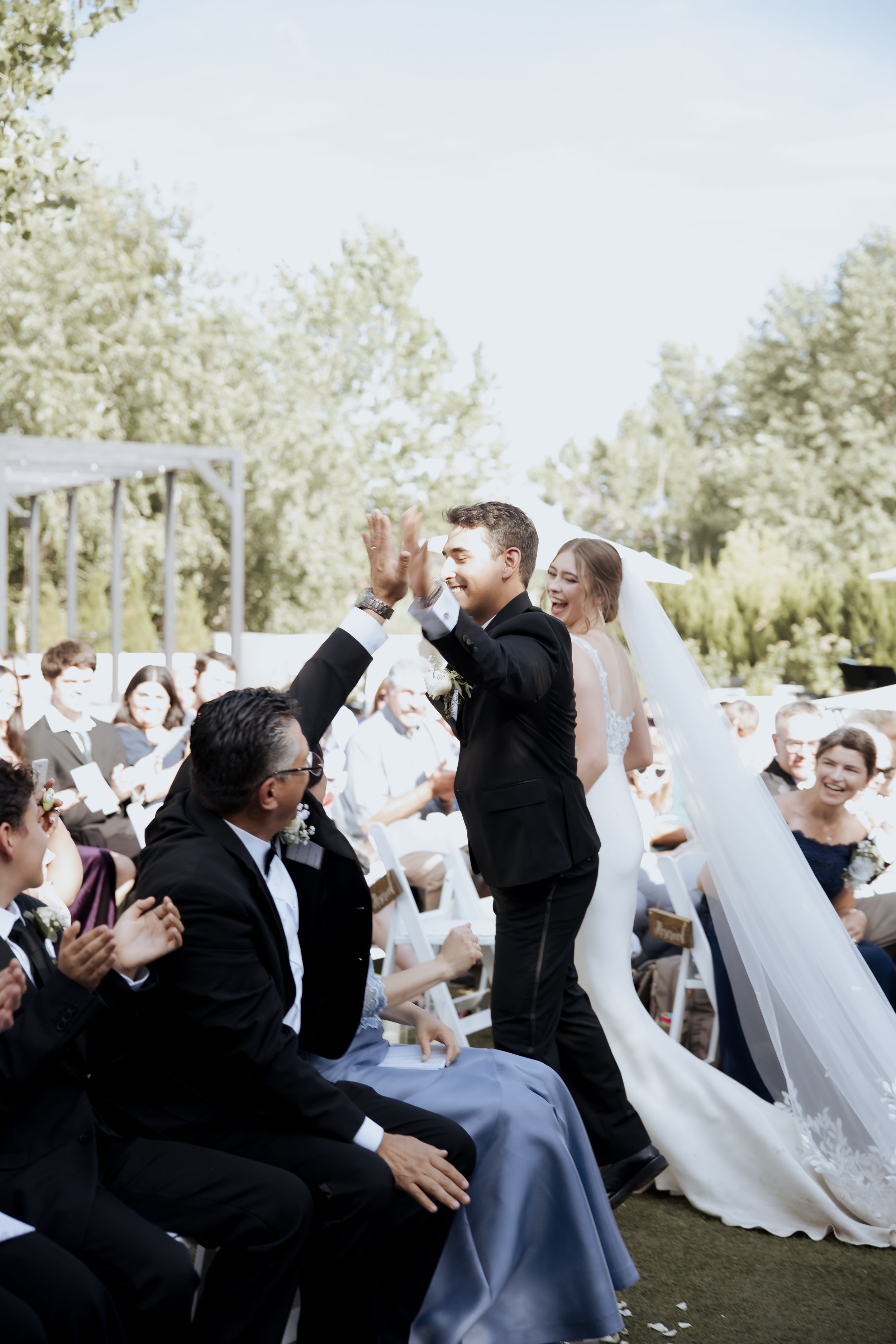 Wedding guests seated outdoors celebrate with raised arms during a joyful celebration moment.