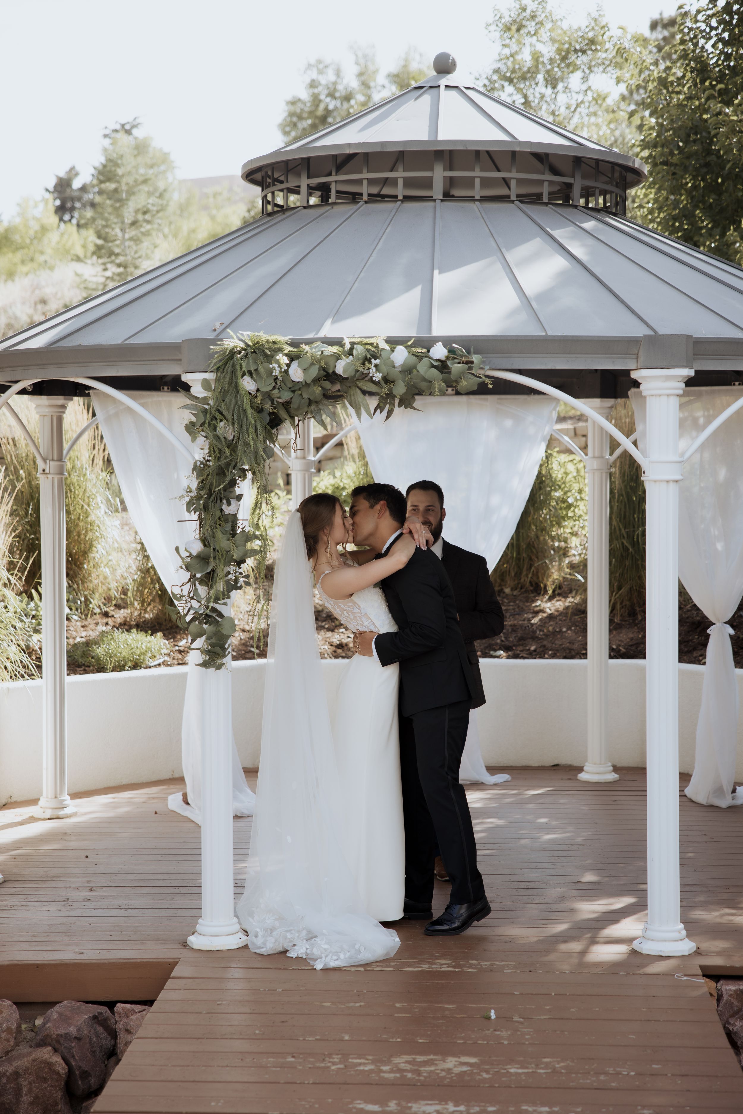 Elegant outdoor wedding ceremony under a decorated gazebo with white drapes and floral arrangements on a wooden deck.