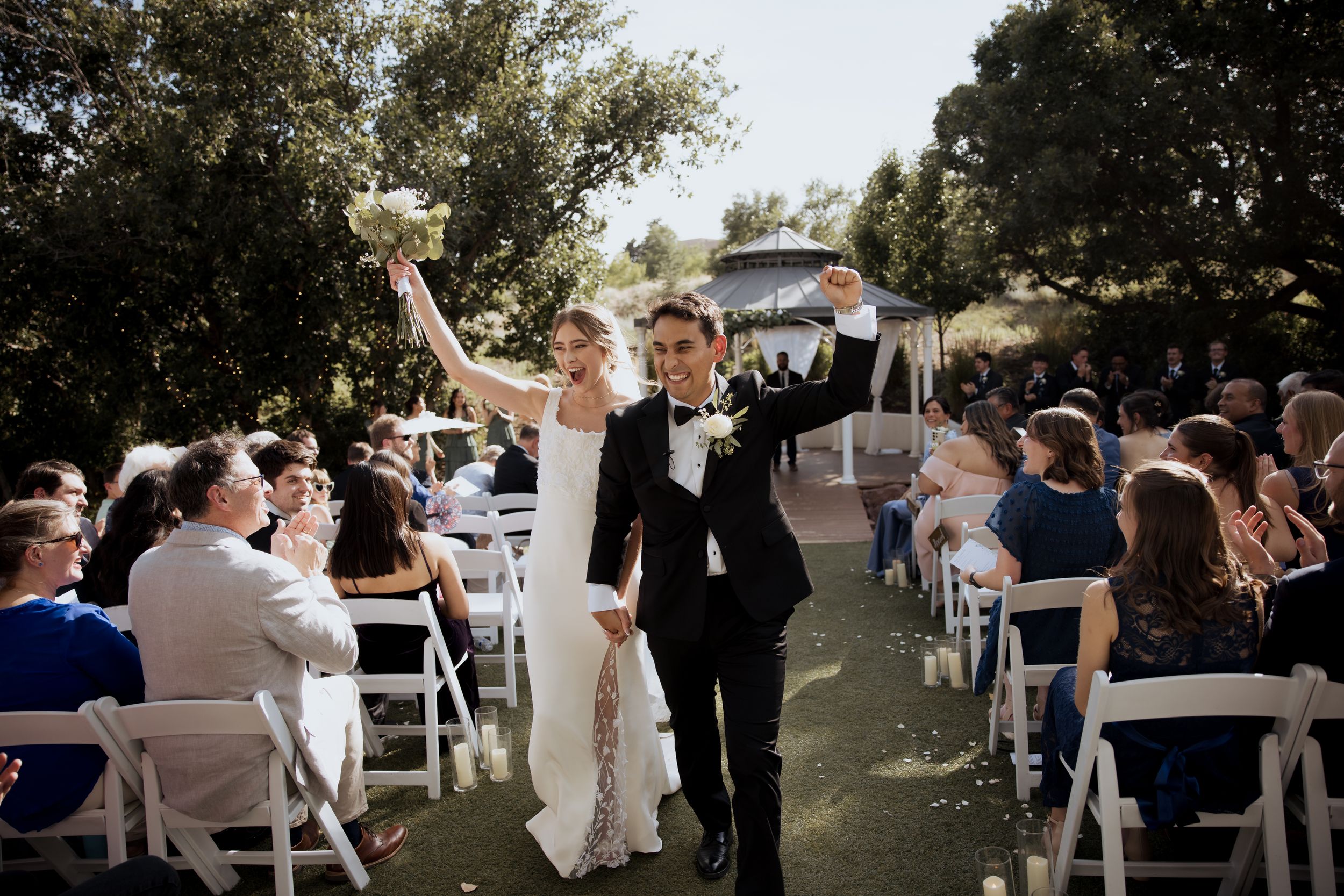 A joyful couple celebrates while walking down the aisle at an outdoor wedding ceremony with guests seated on white chairs.