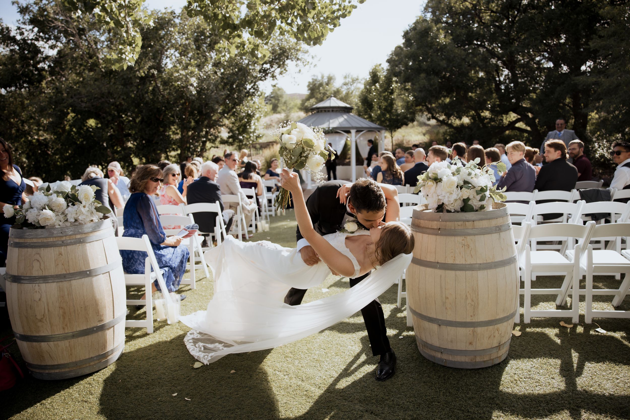 A romantic dip between newlyweds at an outdoor winery wedding ceremony with rustic wine barrels and white chairs.
