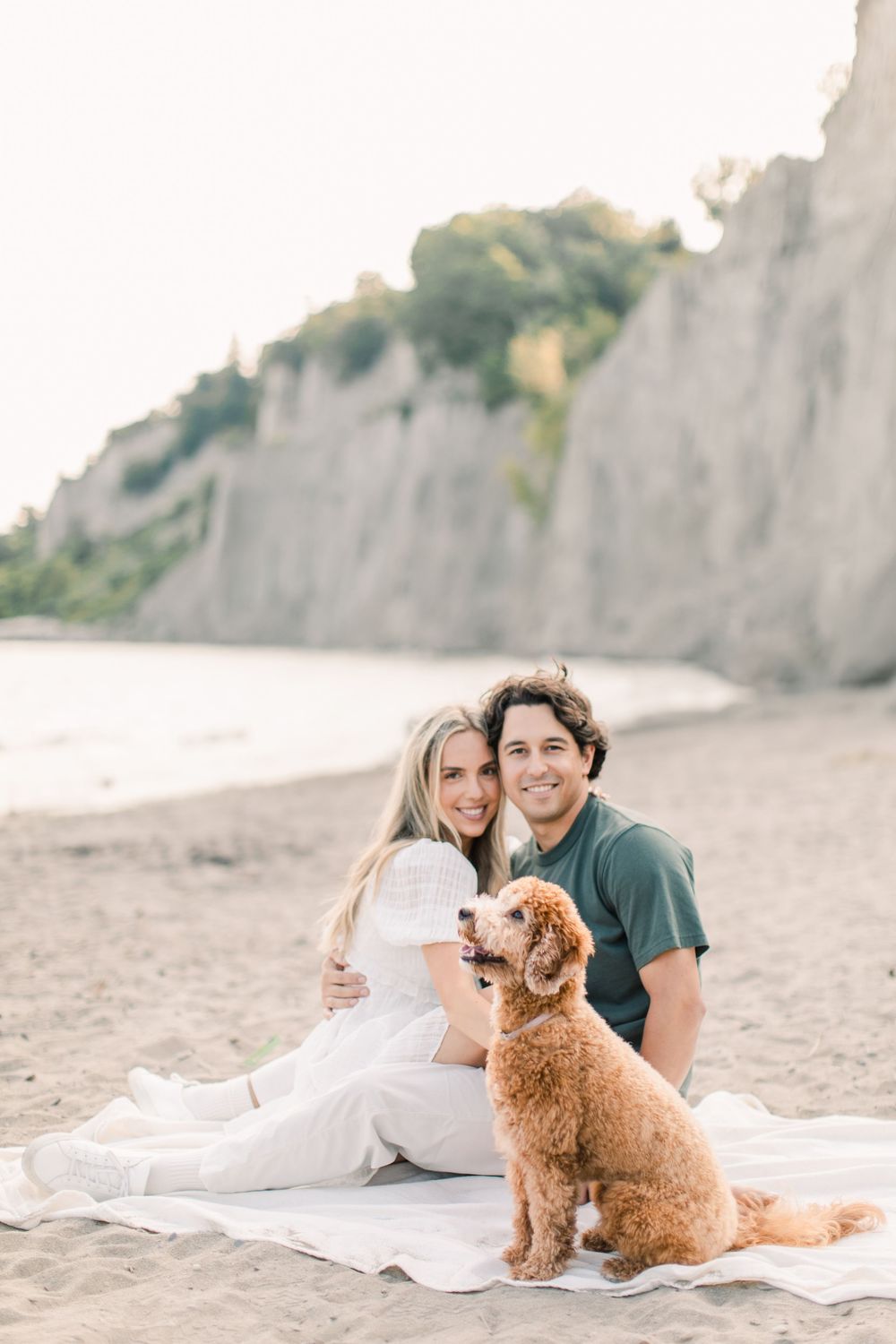 Couple relaxing on beach blanket with their fluffy dog near coastal cliffs.