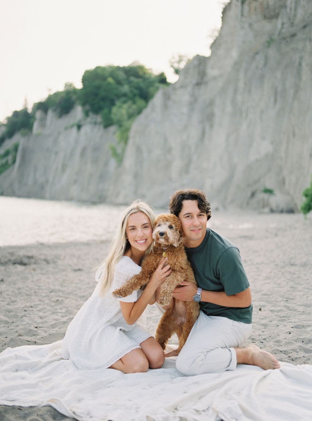 Happy couple cuddling with their goldendoodle on a white blanket at the beach.