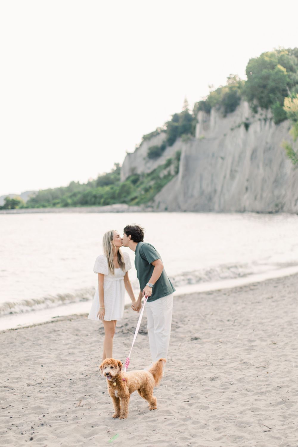 Playful couple on sandy beach with their golden retriever near tall cliffs.