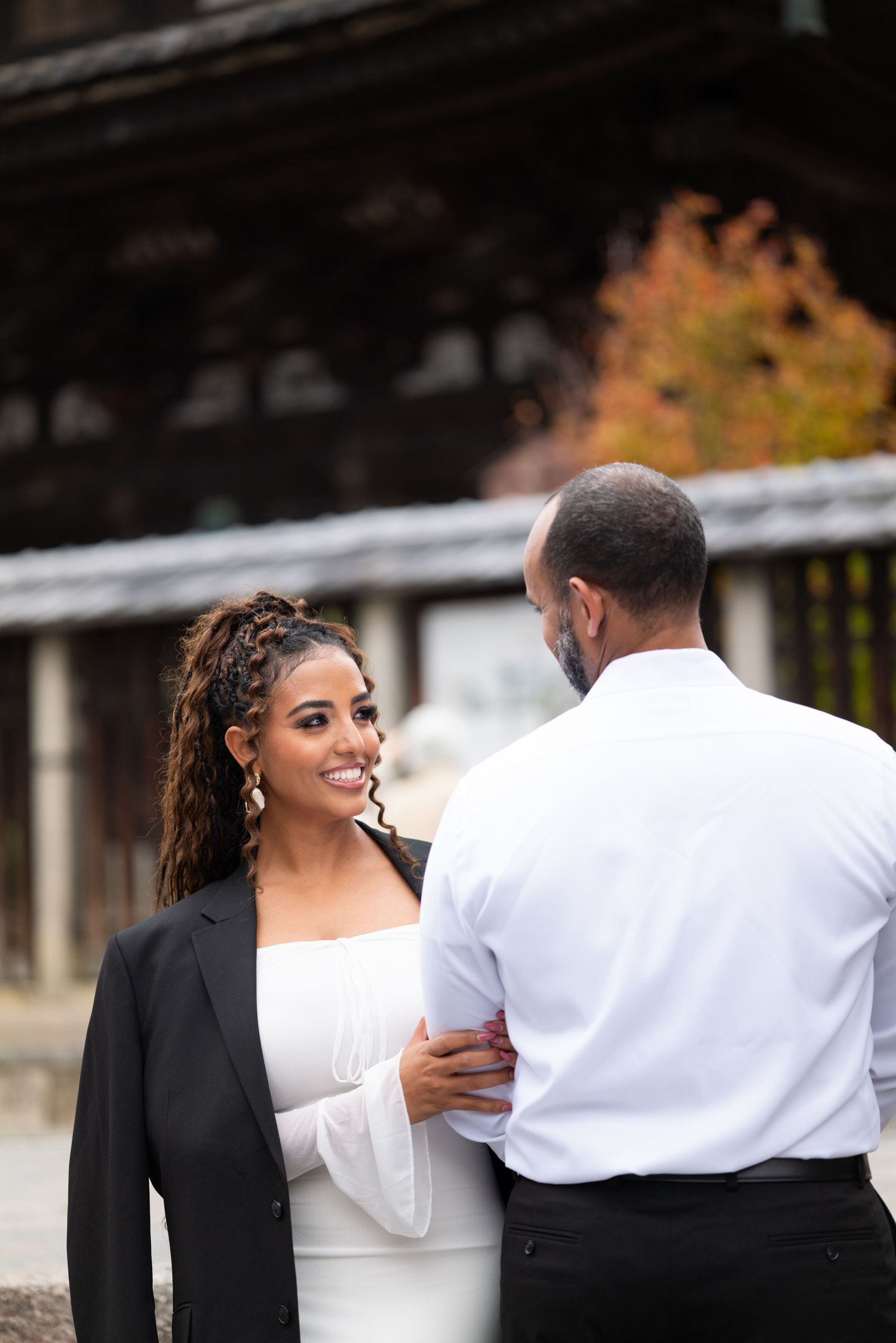 Smiling bride in white dress and black blazer holds groom's arm at a Japanese shrine.