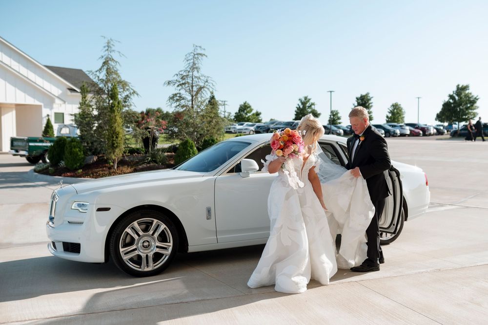 Wedding party members walk through a sunny parking lot near luxury vehicles.