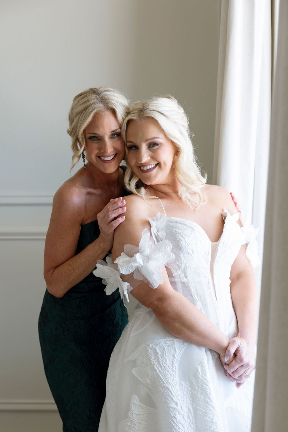 Two women share a joyful moment together, one wearing a white wedding gown and the other in an elegant black dress.