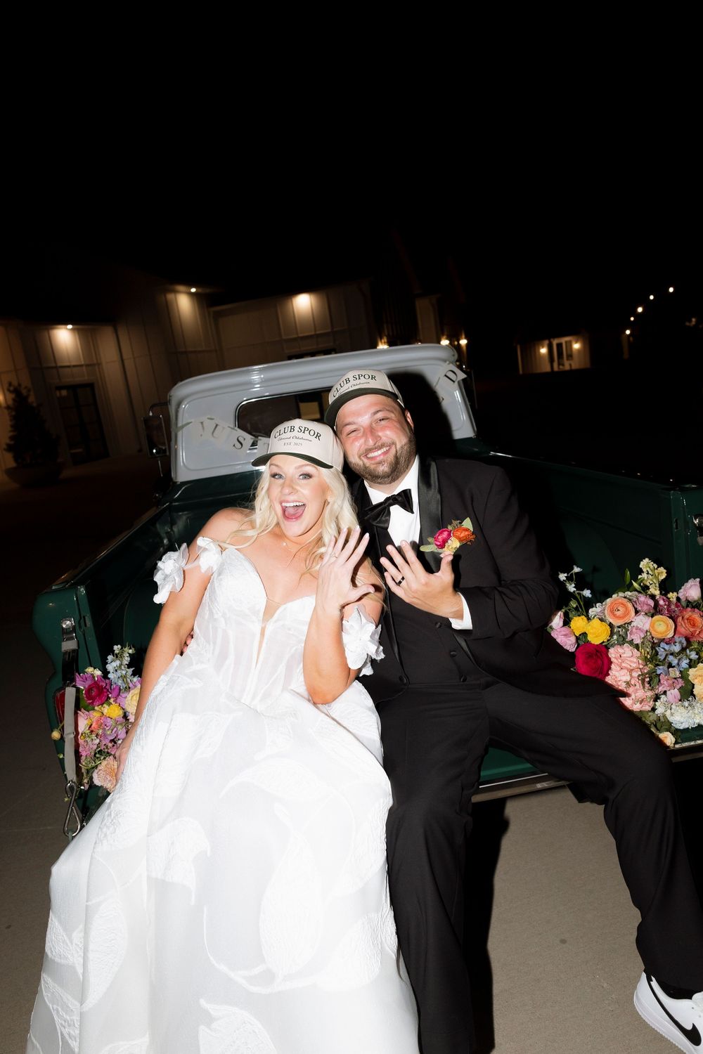 A playful wedding couple poses together at night with colorful floral arrangements in the background.