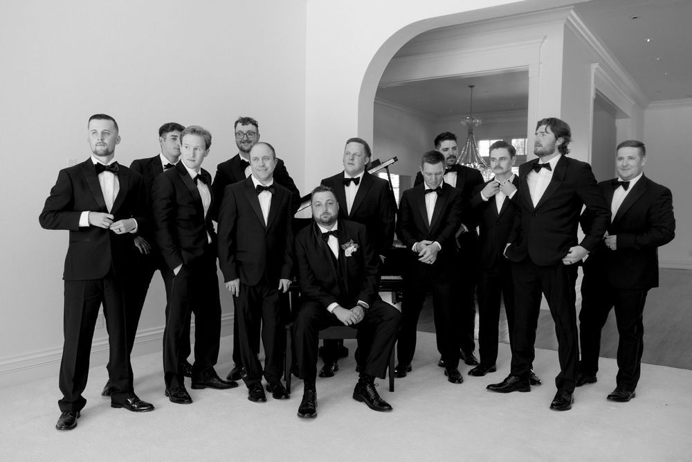 A group of groomsmen in black tuxedos pose together for a formal black and white wedding photo.