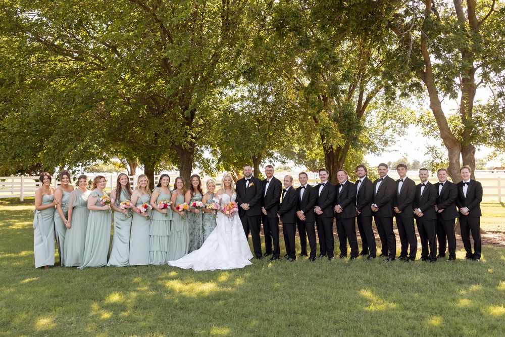Wedding party poses outdoors in a line formation against a backdrop of green trees.