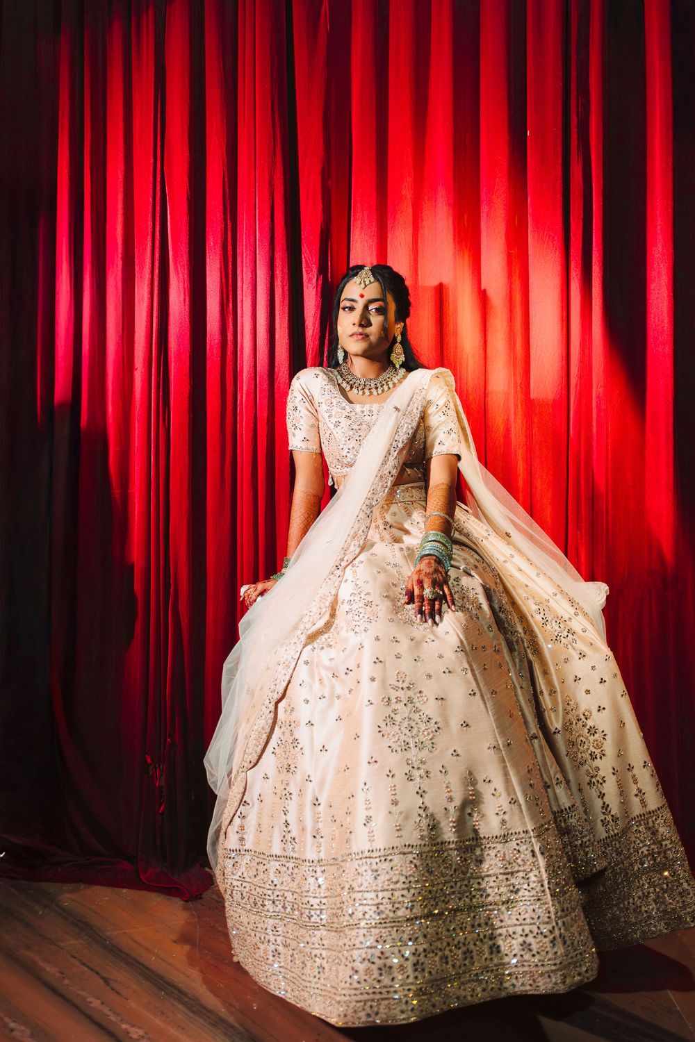 A bride in an ornate ivory lehenga with intricate embroidery poses against a dramatic red curtain backdrop.