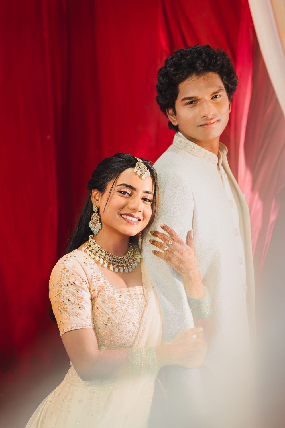 Wedding photoshoot against red curtain backdrop showing couple in traditional Indian attire.
