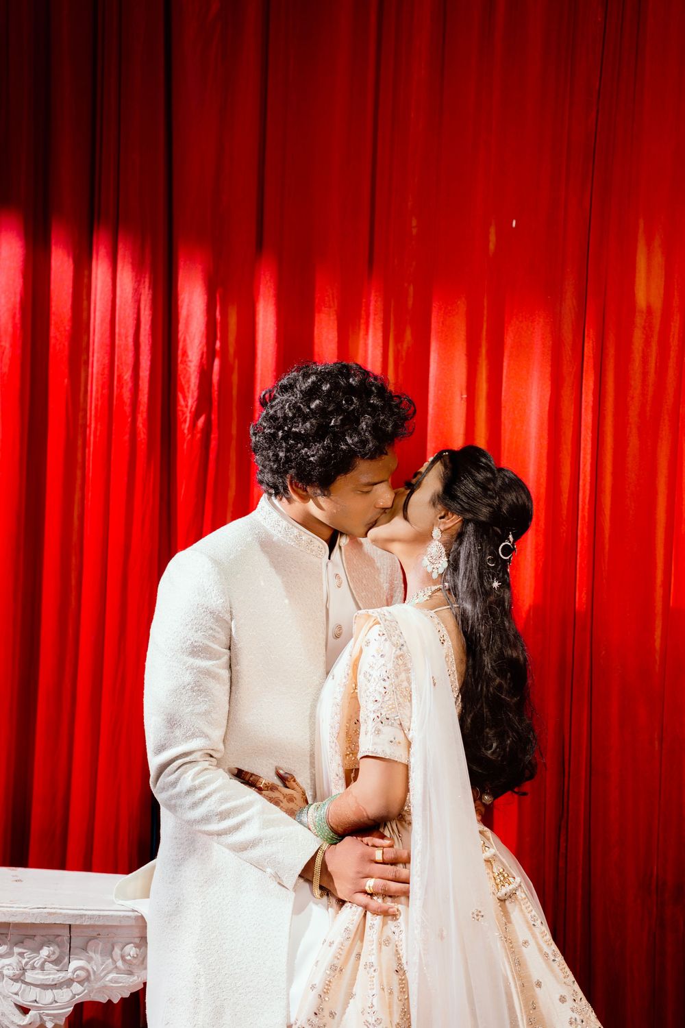 A couple in white attire shares a romantic moment against a dramatic red curtain backdrop.