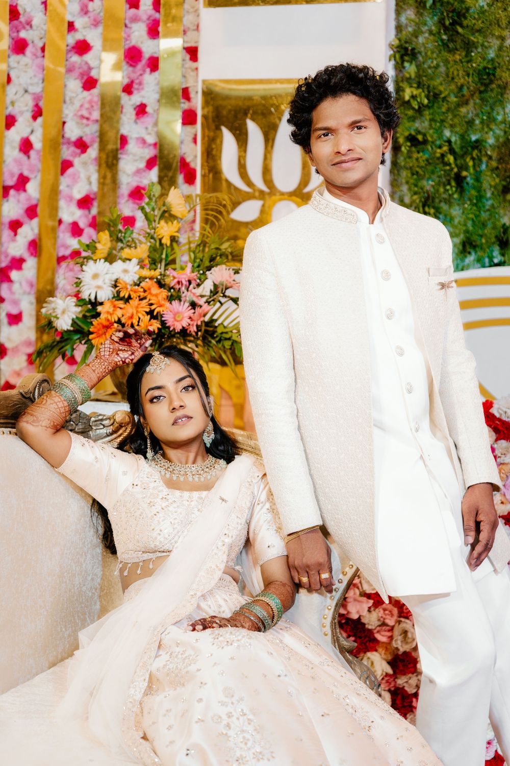 A couple in elegant white attire poses together against a floral backdrop at a formal event.