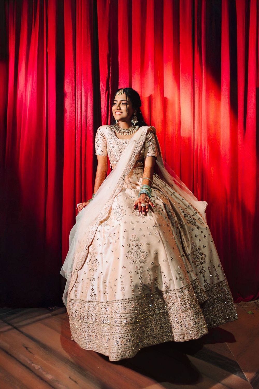 A bride in an ornate white lehenga poses dramatically against a red curtain backdrop in a series of elegant shots.