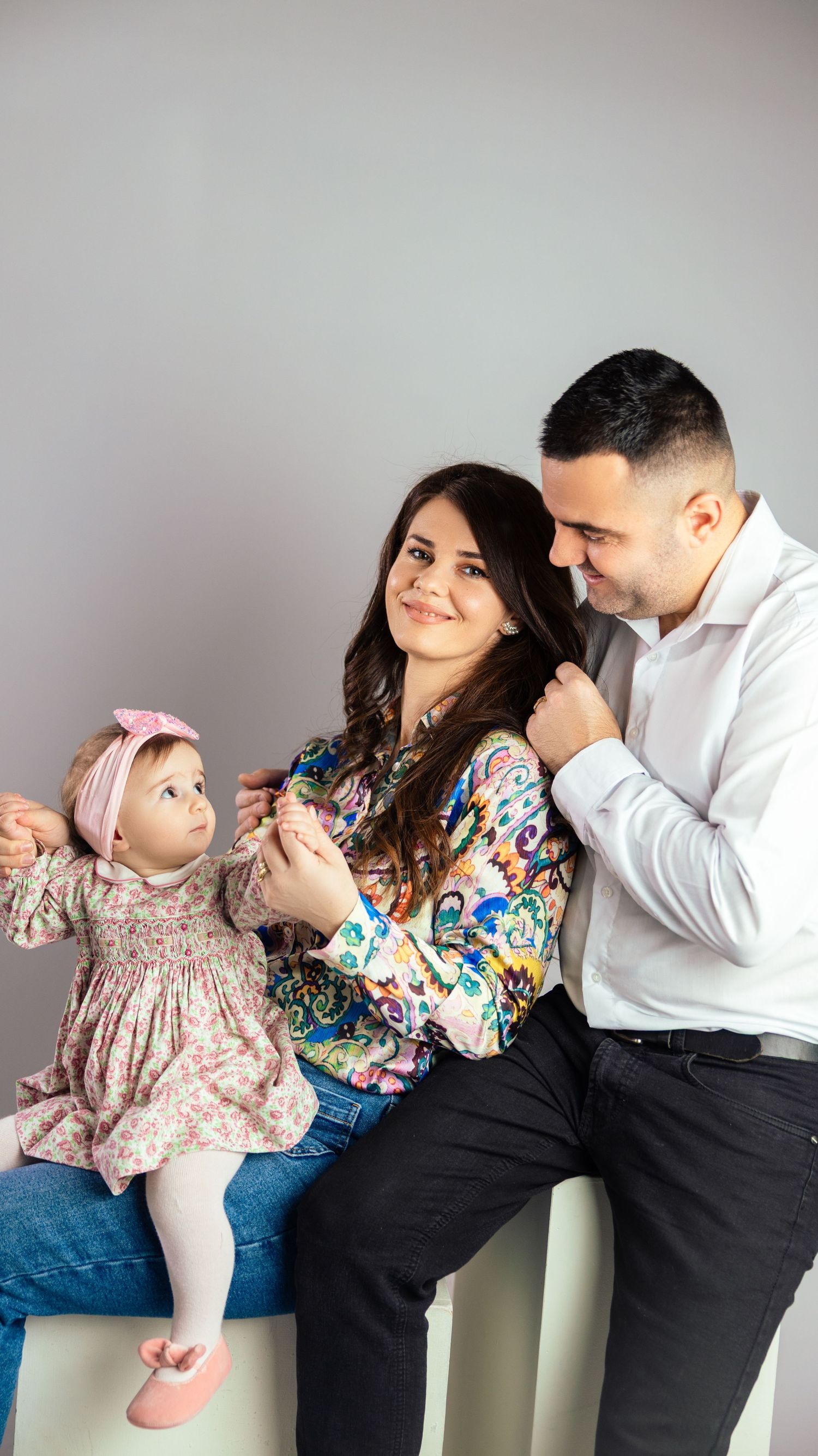 Happy young family with baby girl in floral dress posing together in studio portrait.