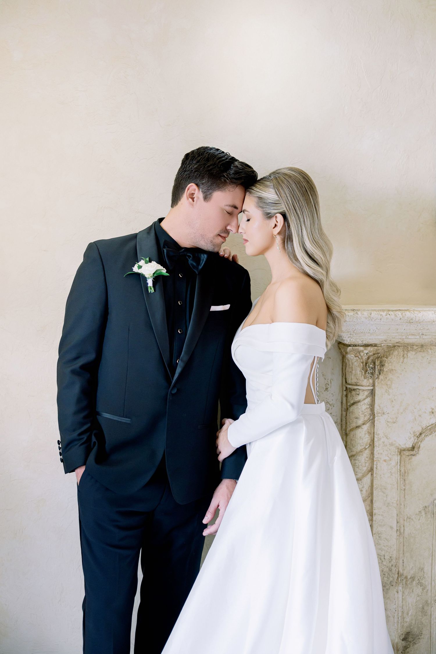 Elegant bride in white strapless gown and groom in dark suit share intimate moment against light neutral wall.