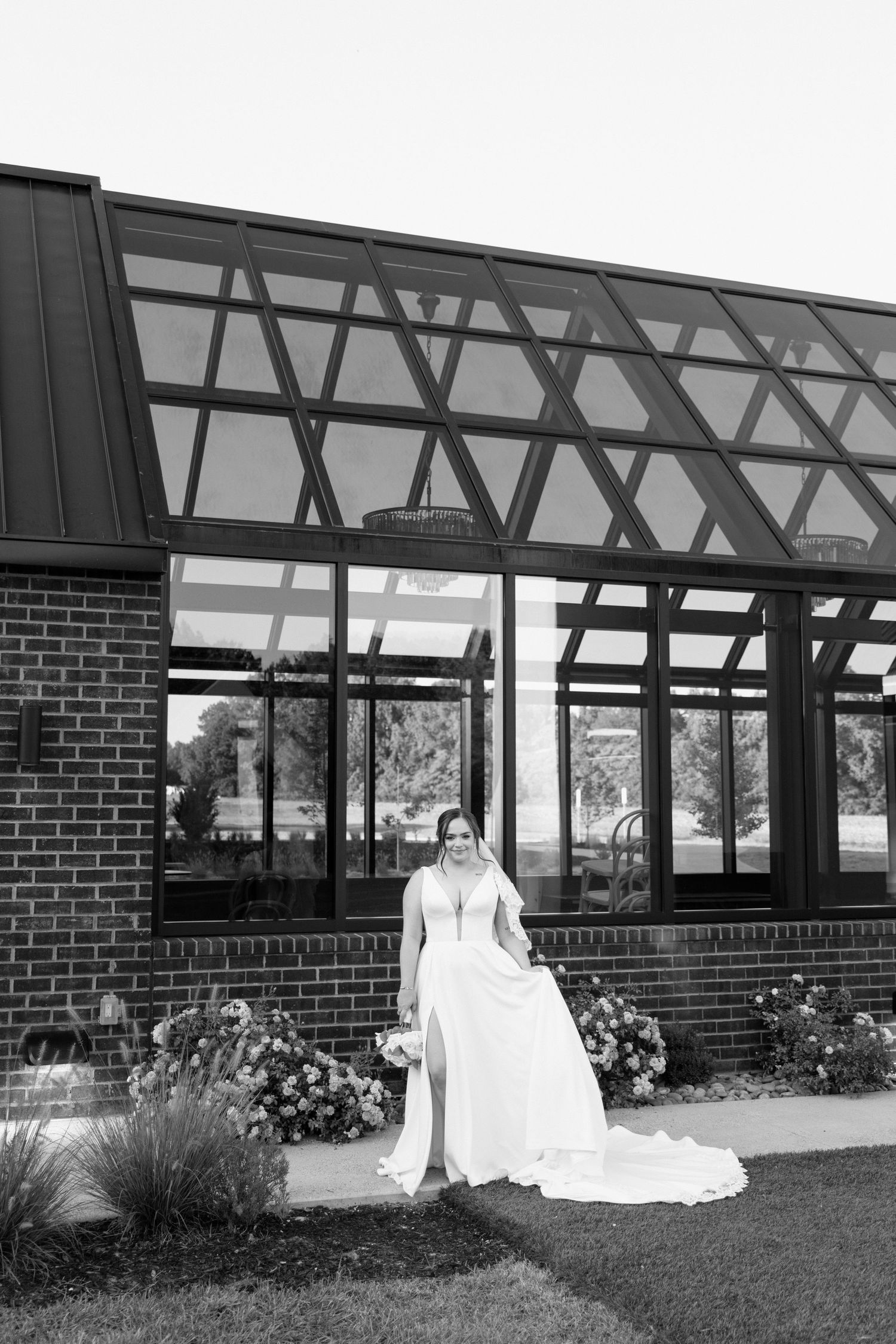 A couple in wedding attire sits together on the grass outside a brick building with large windows.