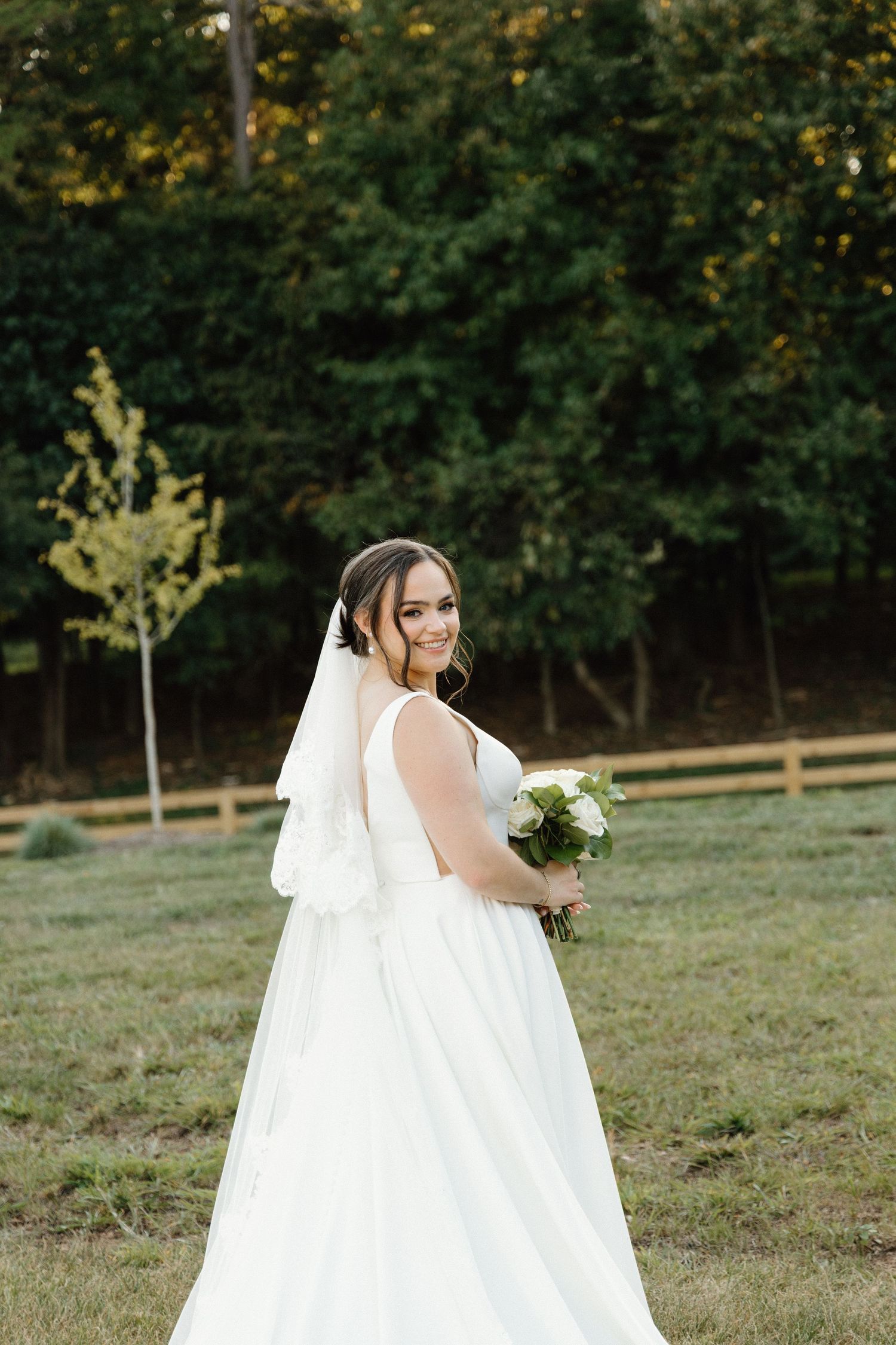 A bride in a flowing white wedding gown and veil poses gracefully in an outdoor setting with trees in the background.