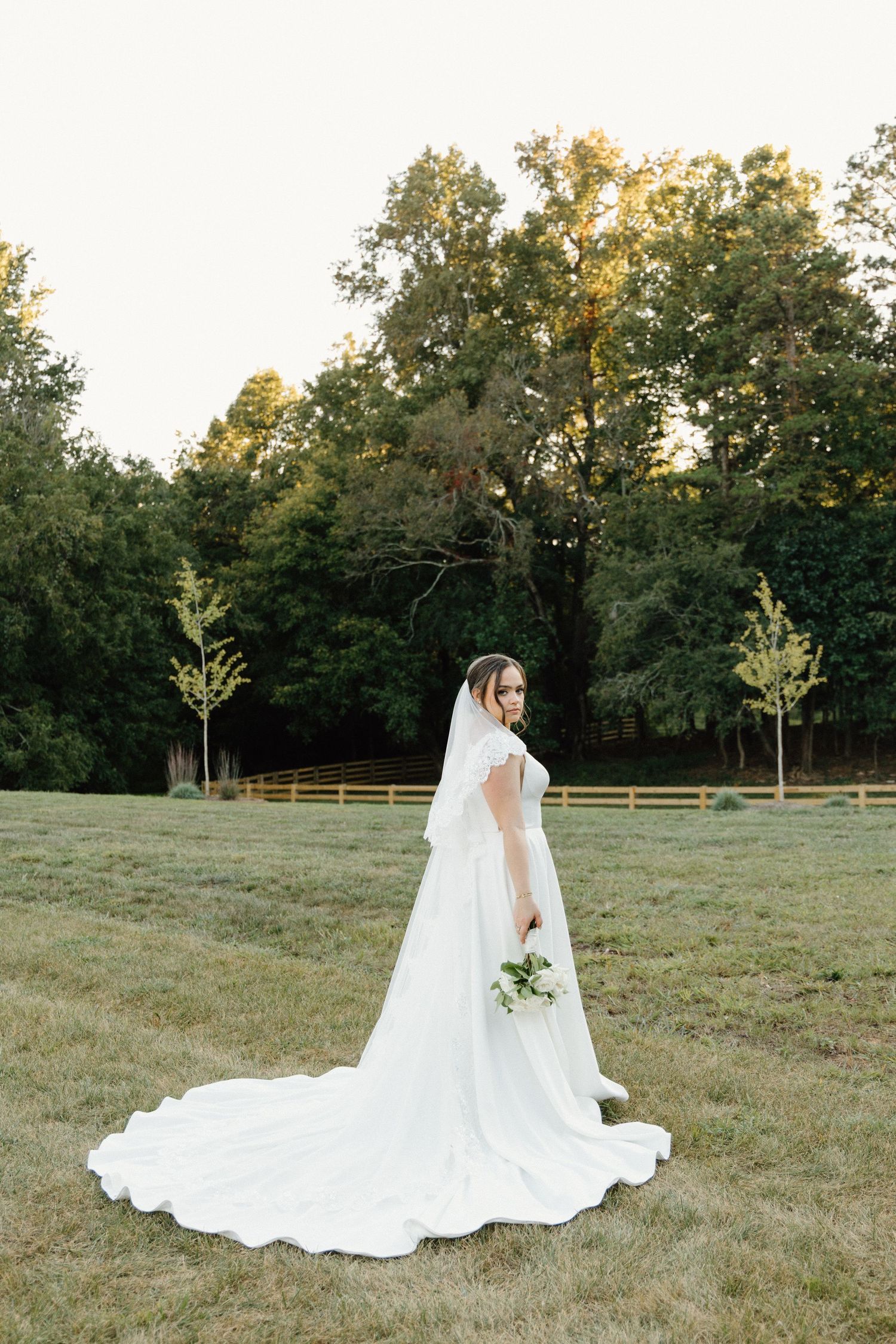 A bride in a flowing white wedding gown with a long cathedral veil poses gracefully in a rustic field at sunset.