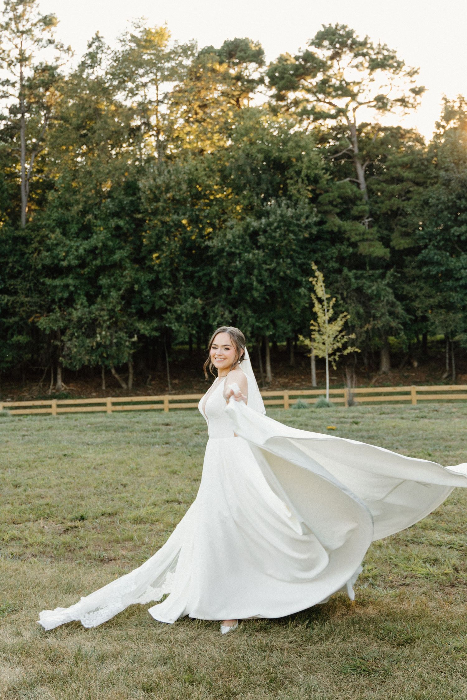 A bride in a flowing white wedding dress twirls gracefully in a grassy field at golden hour.