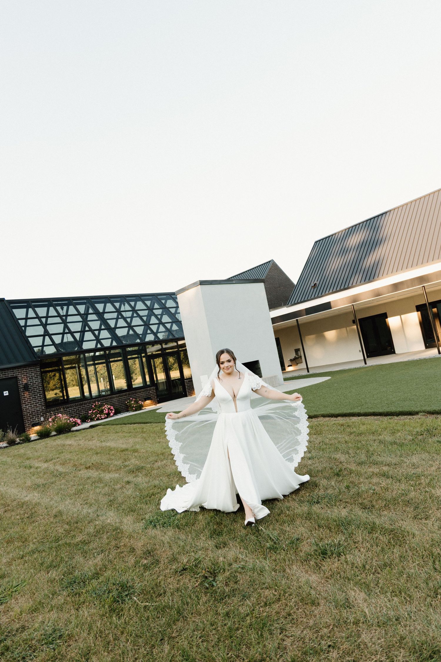 A flowing white wedding dress twirls gracefully across a green lawn with modern architectural buildings in the background.