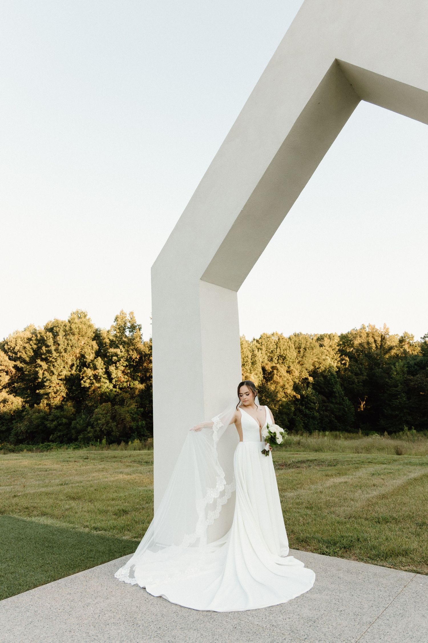 A flowing white wedding dress with a long train creates elegant silhouettes against a natural outdoor backdrop at sunset.