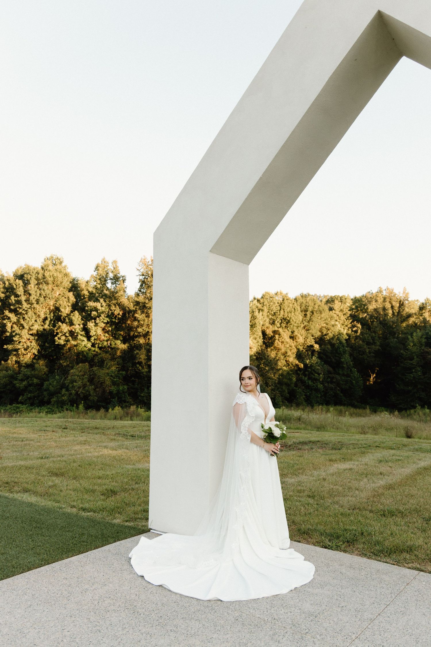 A bride in a flowing white wedding gown poses elegantly against a modern white architectural structure at sunset.