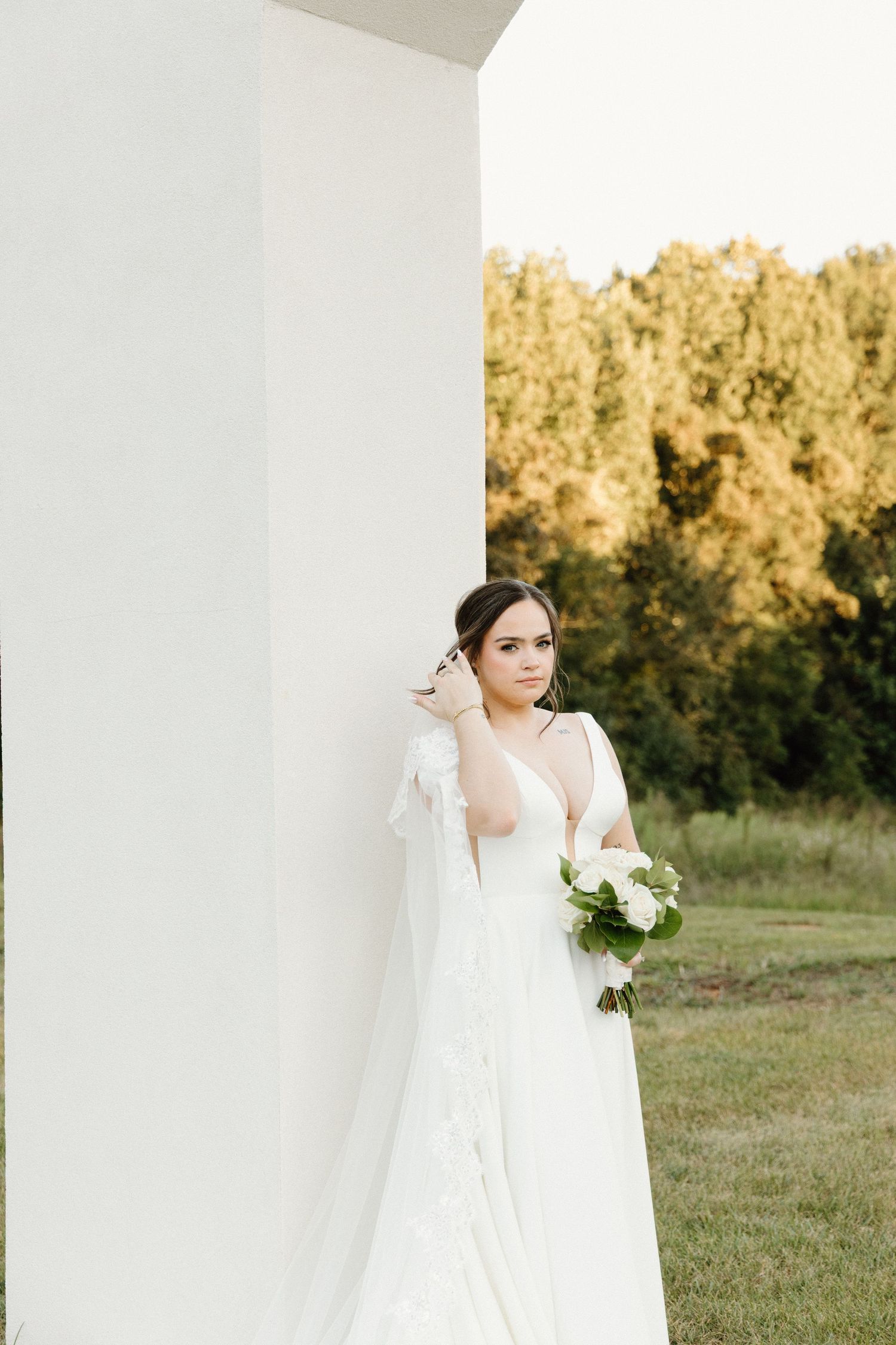 A bride in a flowing white wedding gown stands in a sunlit field with trees in the background during golden hour.