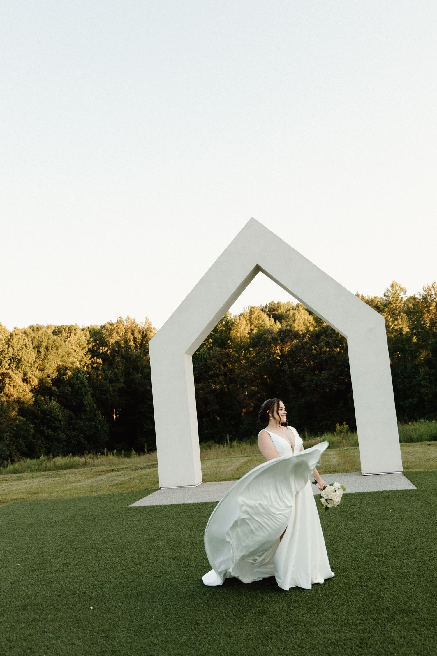 A bride in a flowing white dress twirls gracefully on a green lawn in front of geometric white architectural elements.