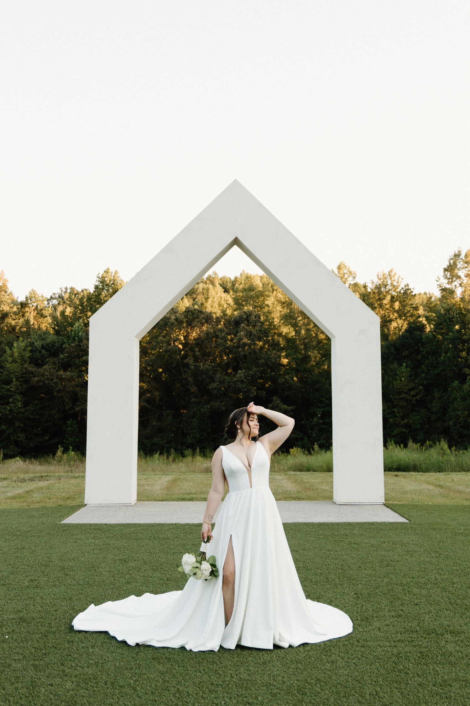 A bride in a flowing white gown poses gracefully against modern white architectural elements on a green lawn at sunset.