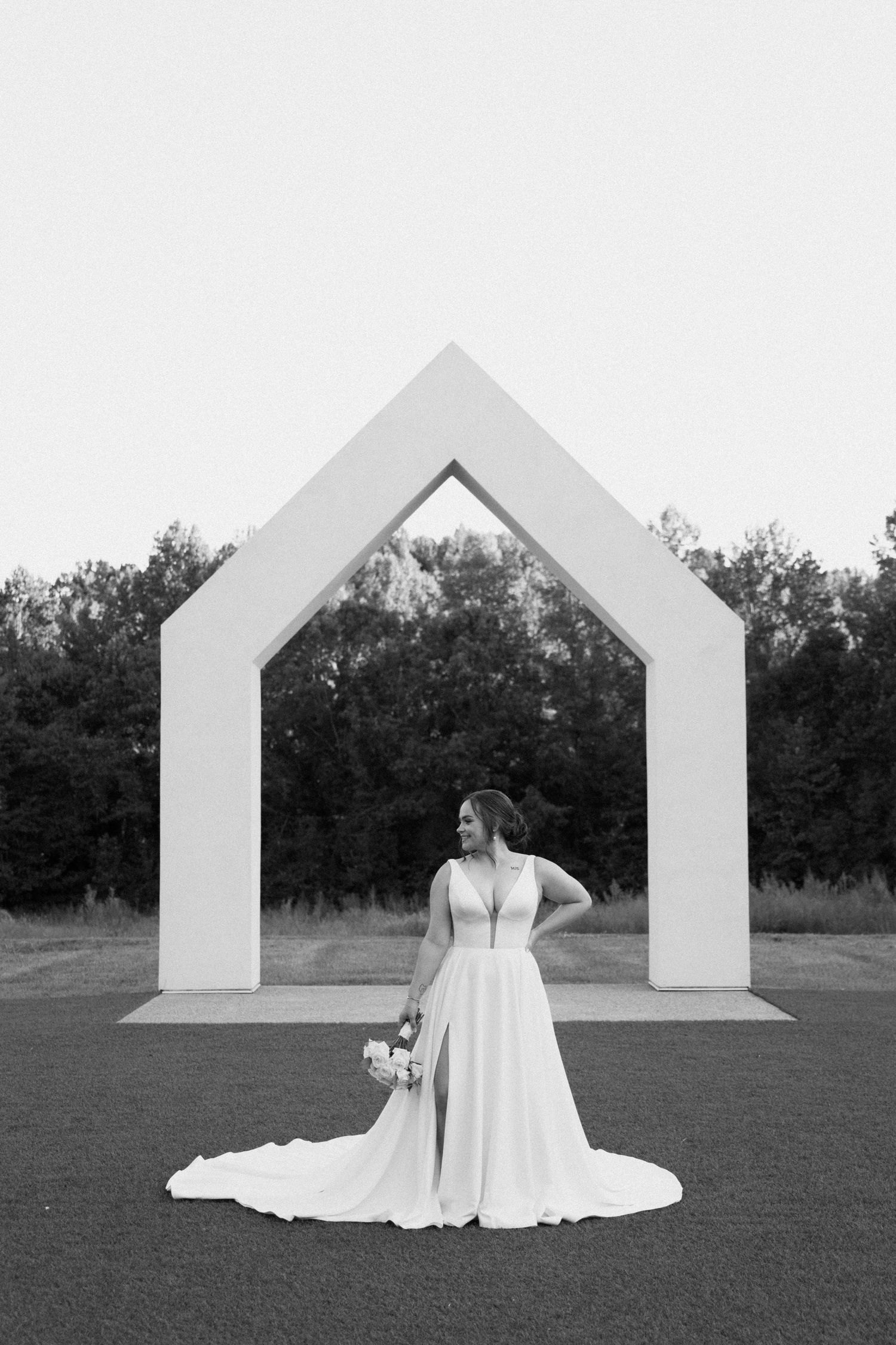 A bride in a flowing white wedding dress stands in front of white geometric house-shaped structures in an outdoor field.