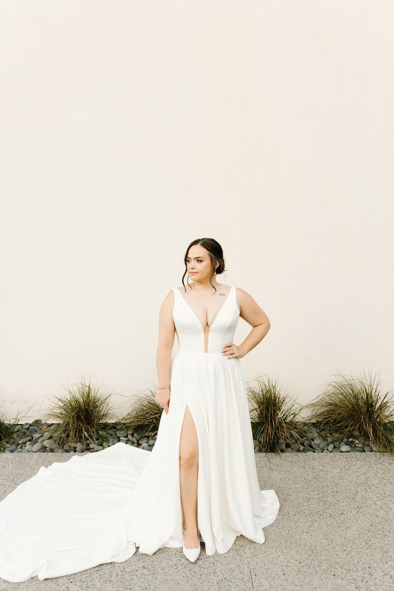 A person in a flowing white dress poses gracefully against a neutral wall with ornamental grasses in an outdoor setting.