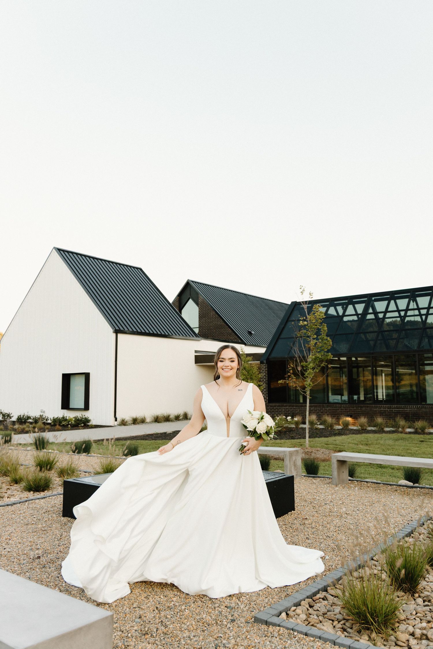 Modern white barn wedding venue with bride twirling in elegant dress on gravel path at sunset.