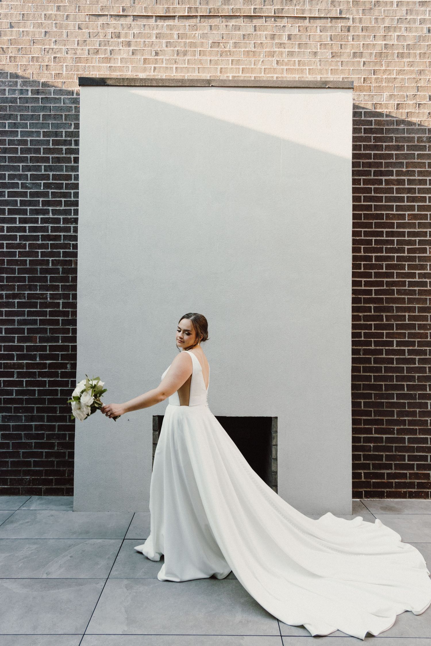 A bride in a flowing white gown with a long train poses against a modern brick and white wall backdrop.