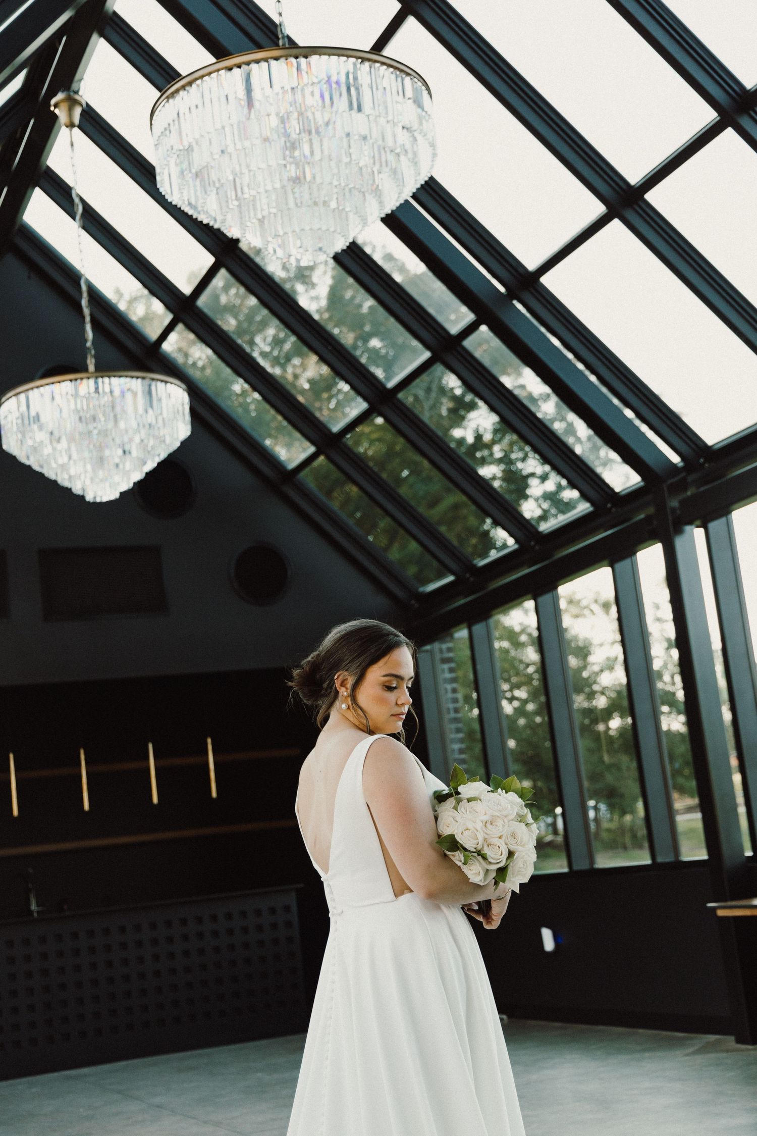 Bride in elegant white wedding dress stands in modern glass atrium with geometric black framing and natural light.