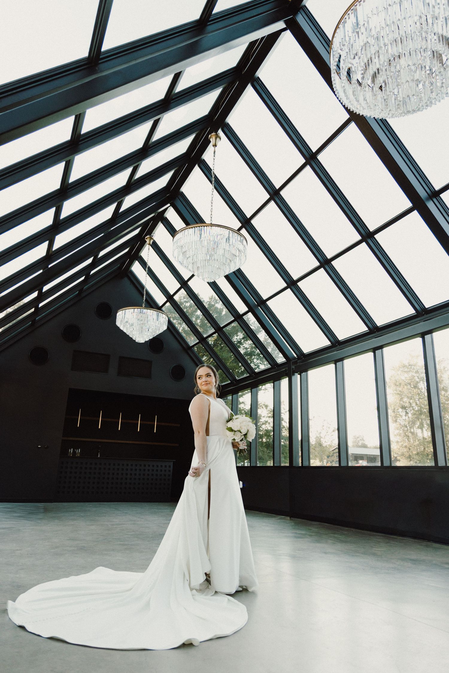 Elegant bride poses in flowing white wedding gown inside modern architectural space with angular glass ceiling and dark walls.
